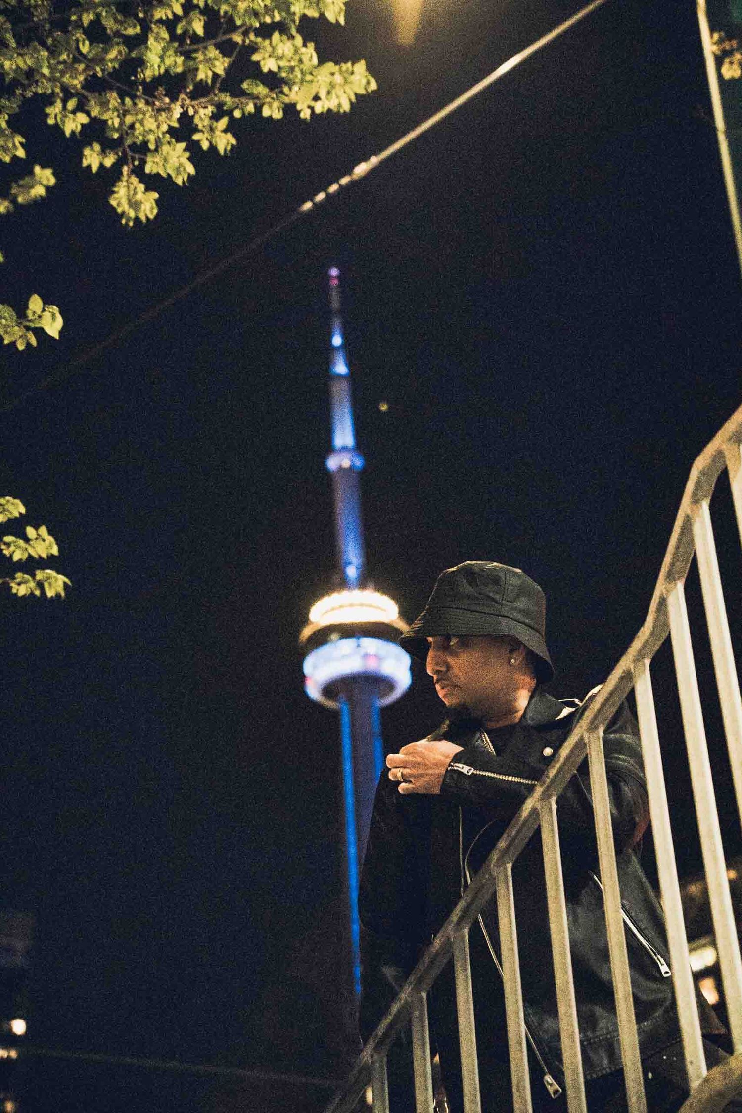 A person in a bucket hat leans on a railing at night with the illuminated CN Tower visible in the background.