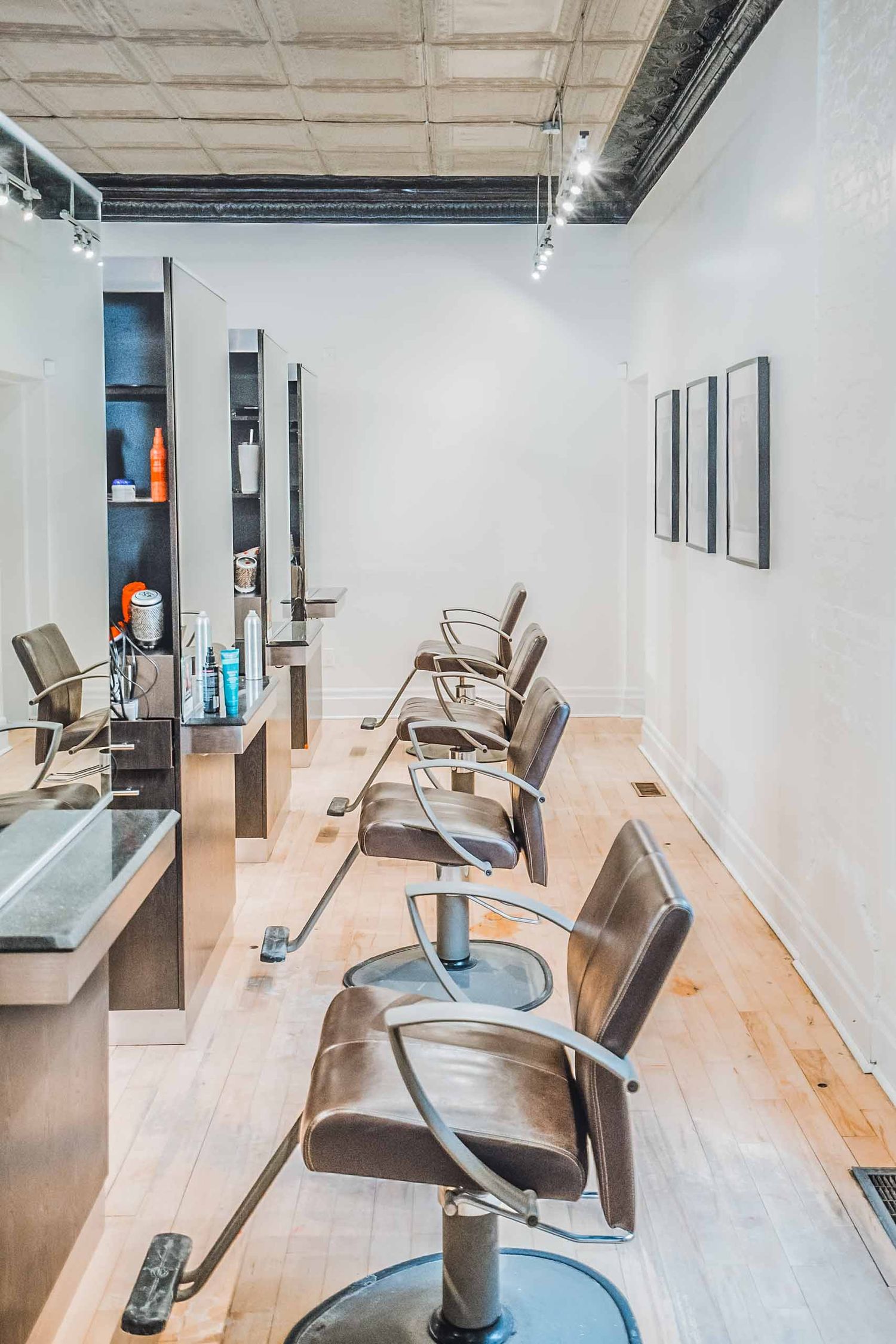 Modern hair salon interior with a row of sleek styling chairs, wood floors, white walls, and product shelving under industrial ceiling.