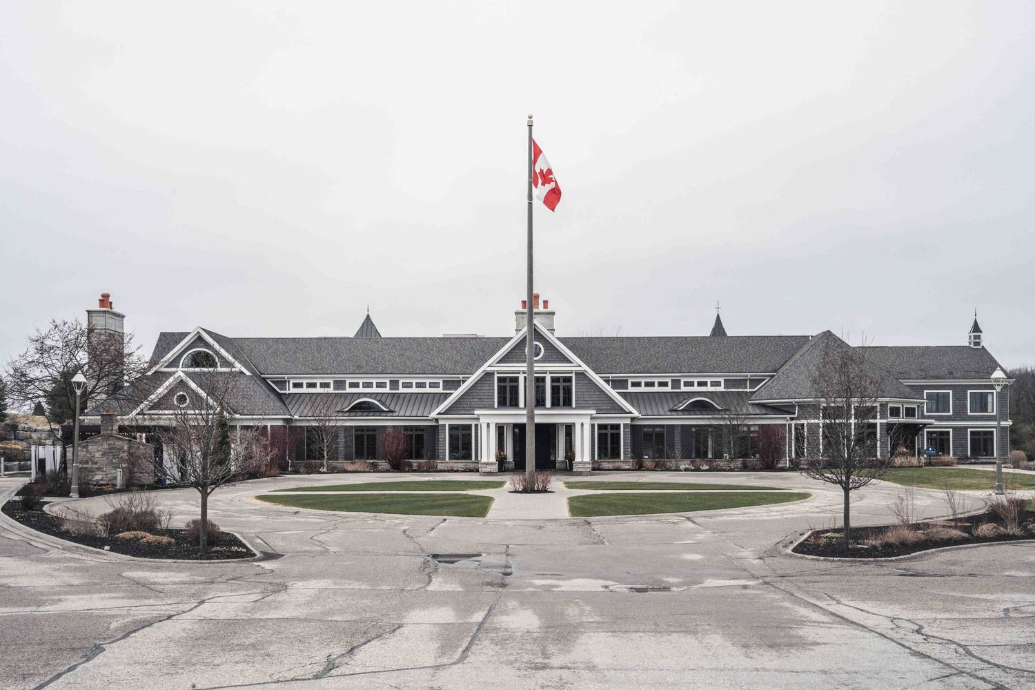 Large Canadian clubhouse with a circular driveway, Canadian flag, and manicured grounds under an overcast sky.