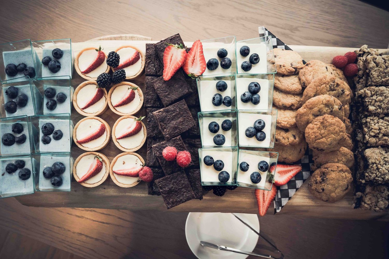 Overhead view of a dessert board with blueberry cheesecakes, tarts, brownies, and oatmeal cookies on a wooden platter.