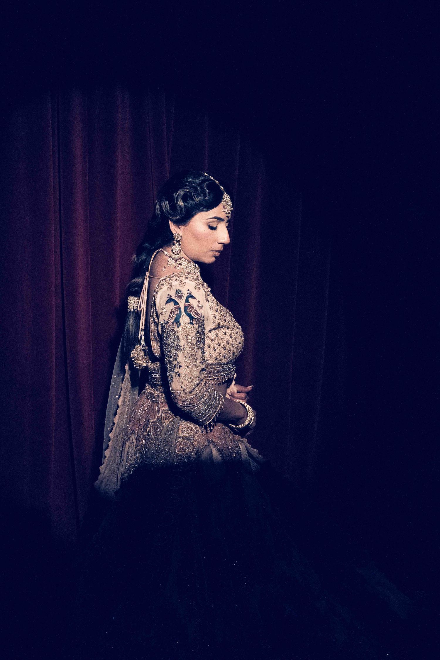 A person in an ornate beige lace dress poses against a dark red curtain backdrop in dramatic lighting.