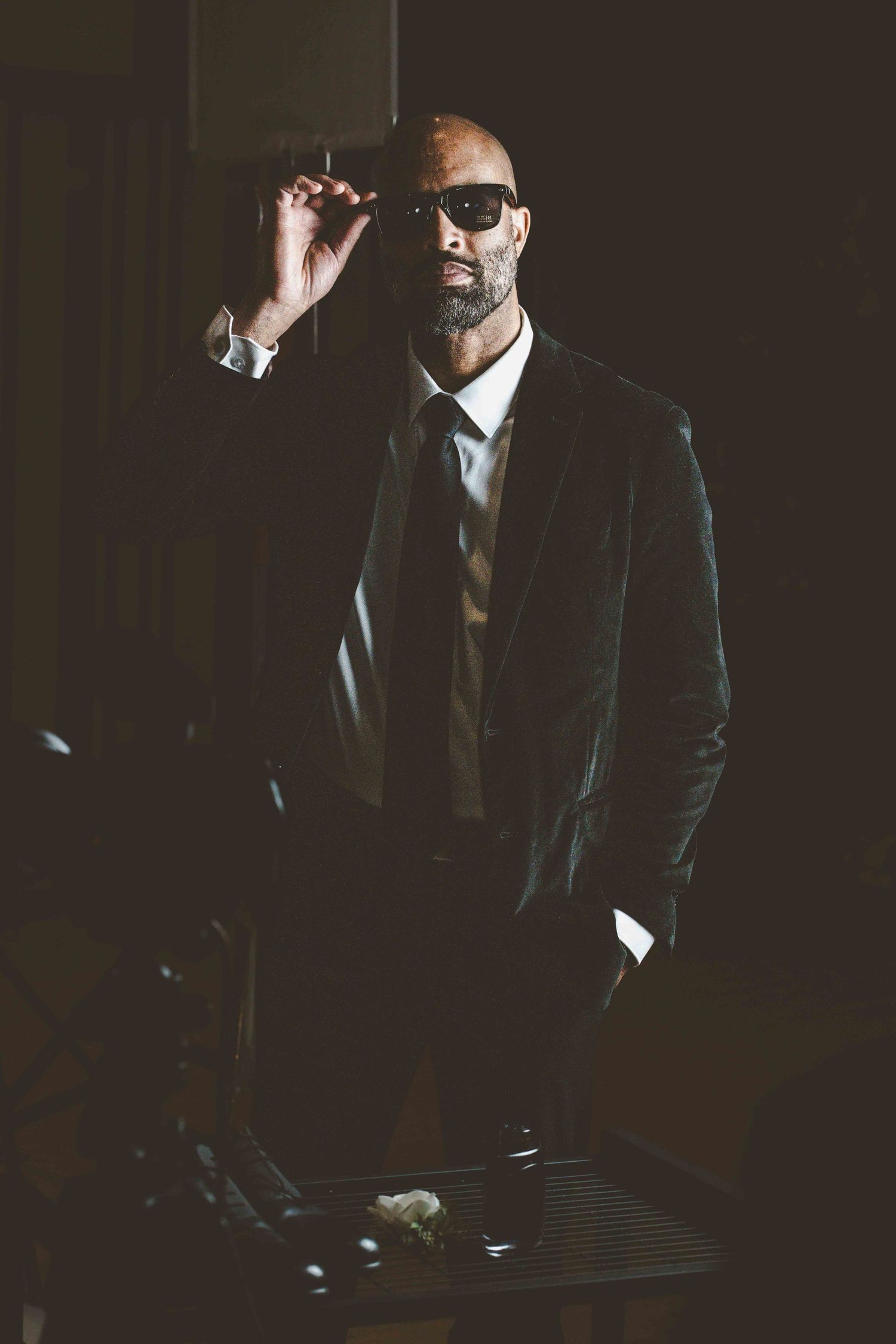 A person in a dark suit and sunglasses poses dramatically in a moody, low-lit environment.
