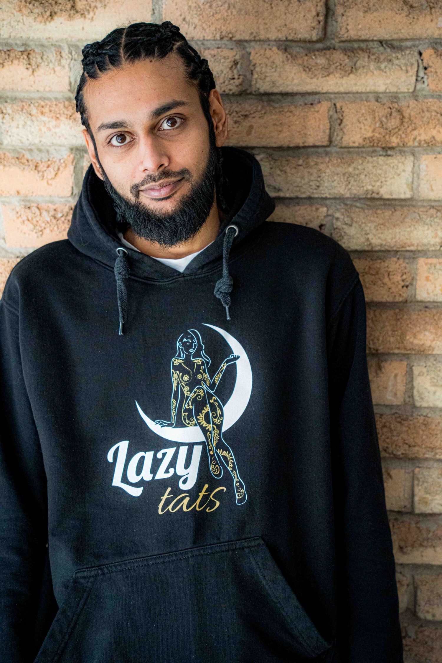 Person wearing black hoodie with moon and text design against brick wall background.