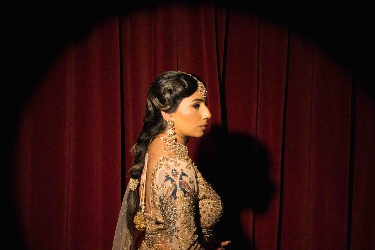 A woman in an ornate gold dress poses in profile against a red curtain backdrop with dramatic lighting.