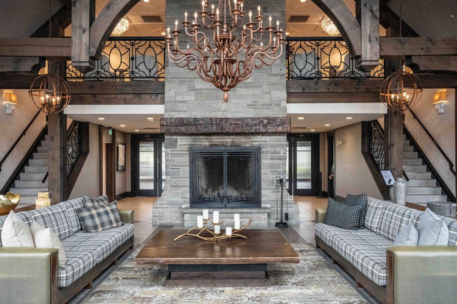 Rustic luxury living room with stone fireplace, plaid sofas, wood beams, chandelier, and dual staircases.
