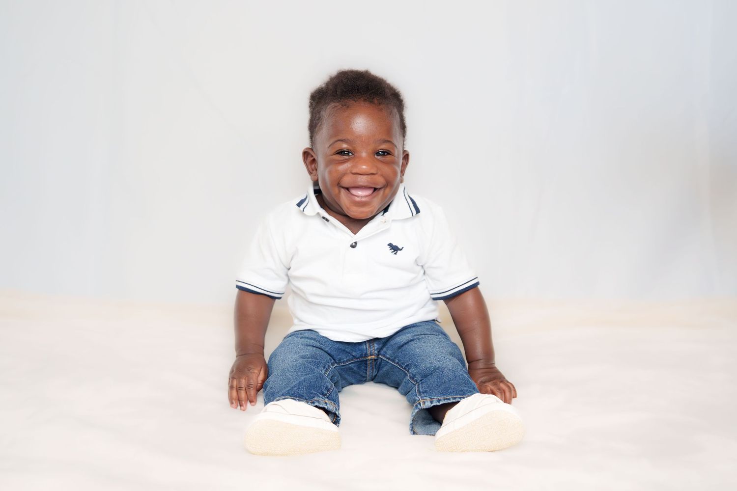 Adorable baby wearing white polo shirt and jeans sitting against light background.