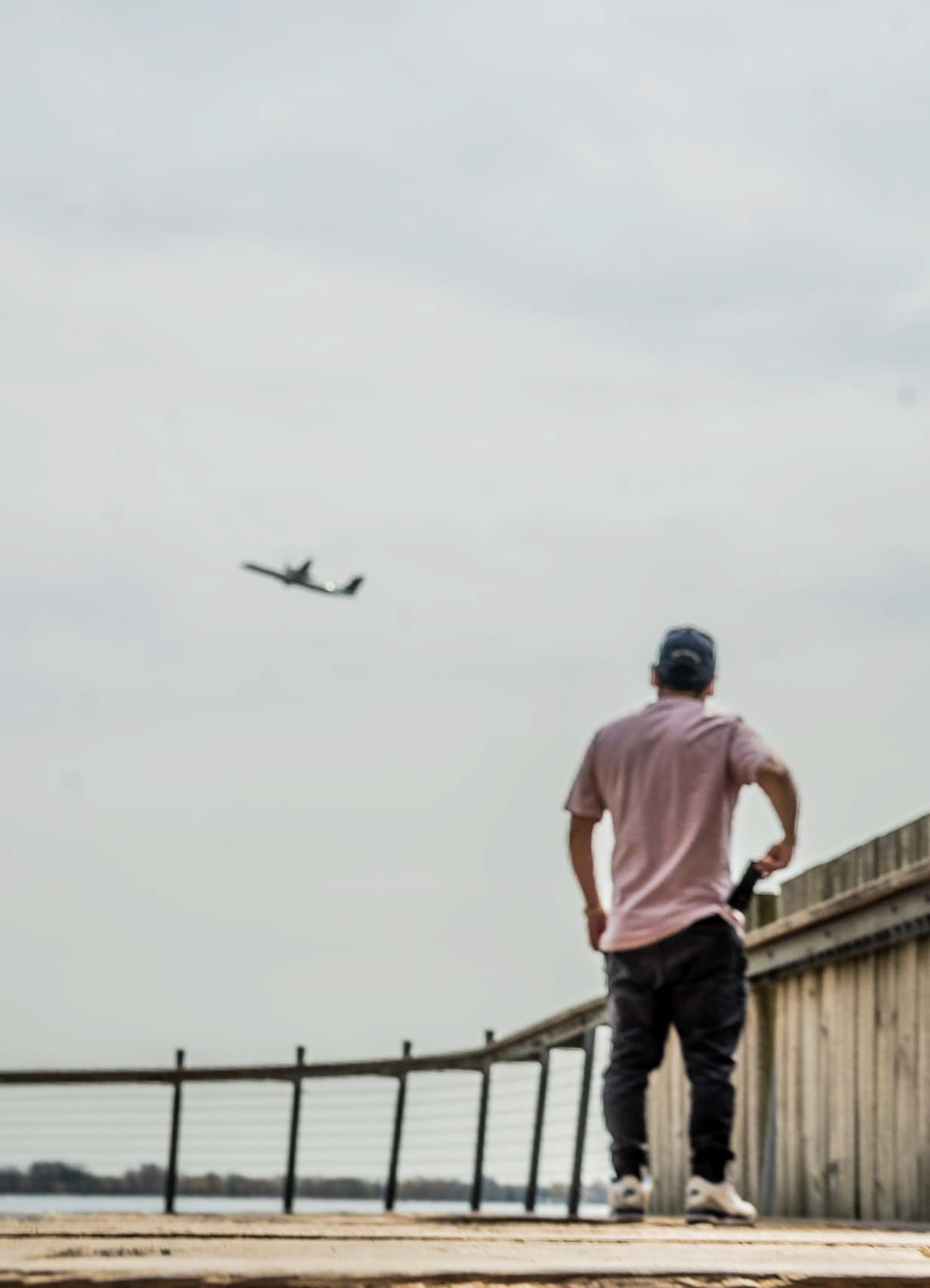Person sitting on edge of wooden pier overlooking water with airplane flying in distance.