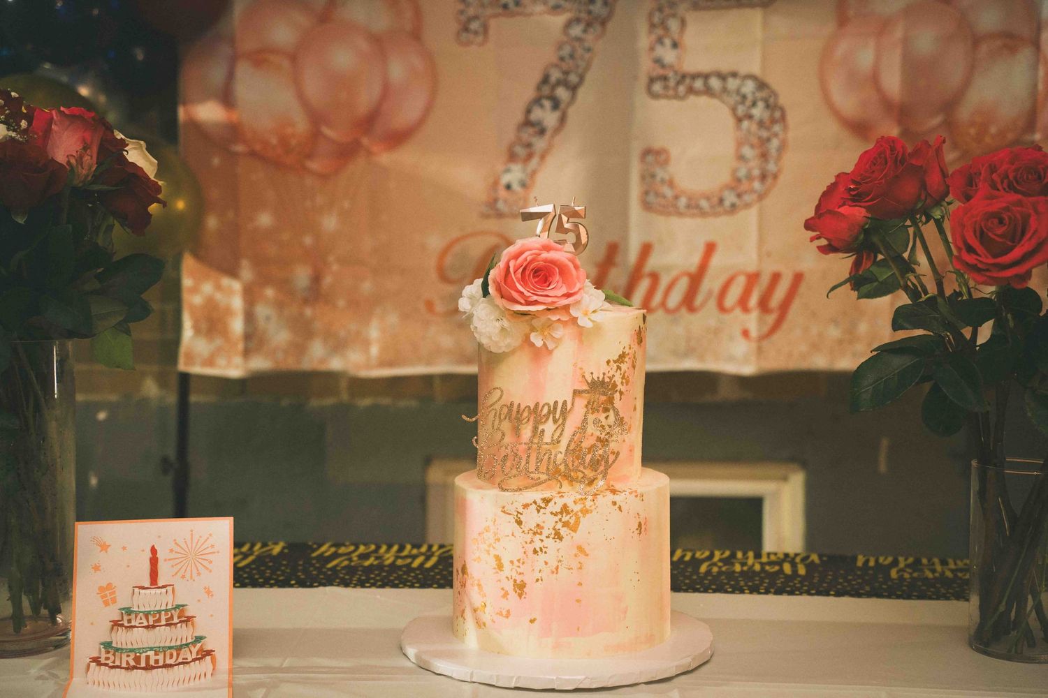 Two-tiered white birthday cake adorned with pink roses and metallic gold embellishments.