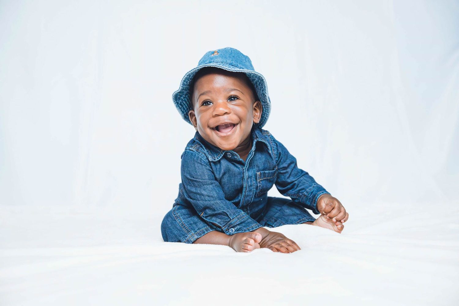 Baby wearing denim outfit and blue hat lying on white background.