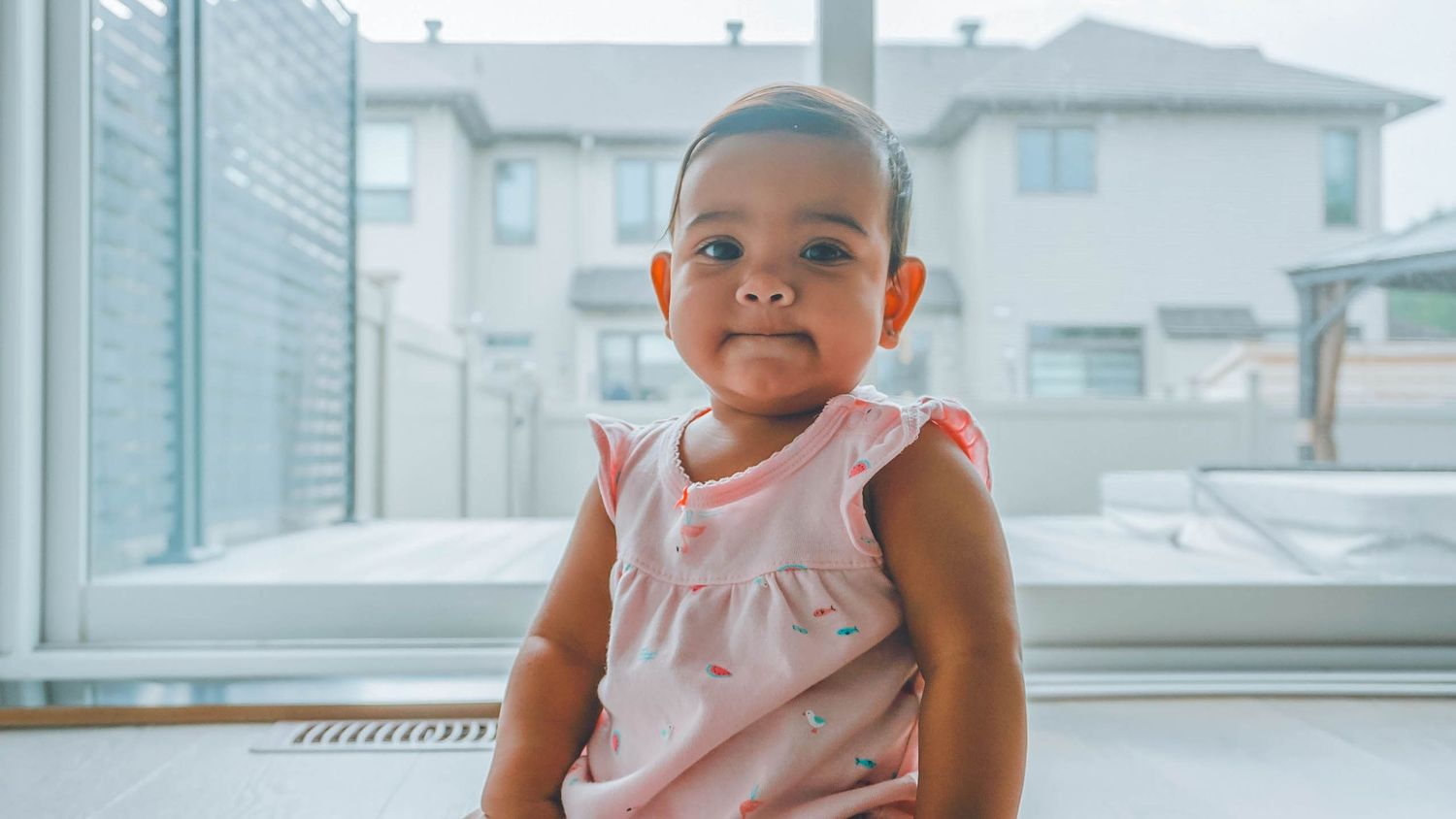 Baby in pink dress sitting by window in bright room.