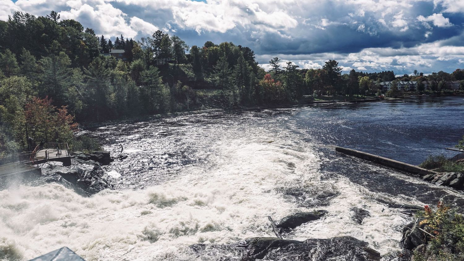 Dramatic panorama of rushing river rapids with white water against cloudy sky.