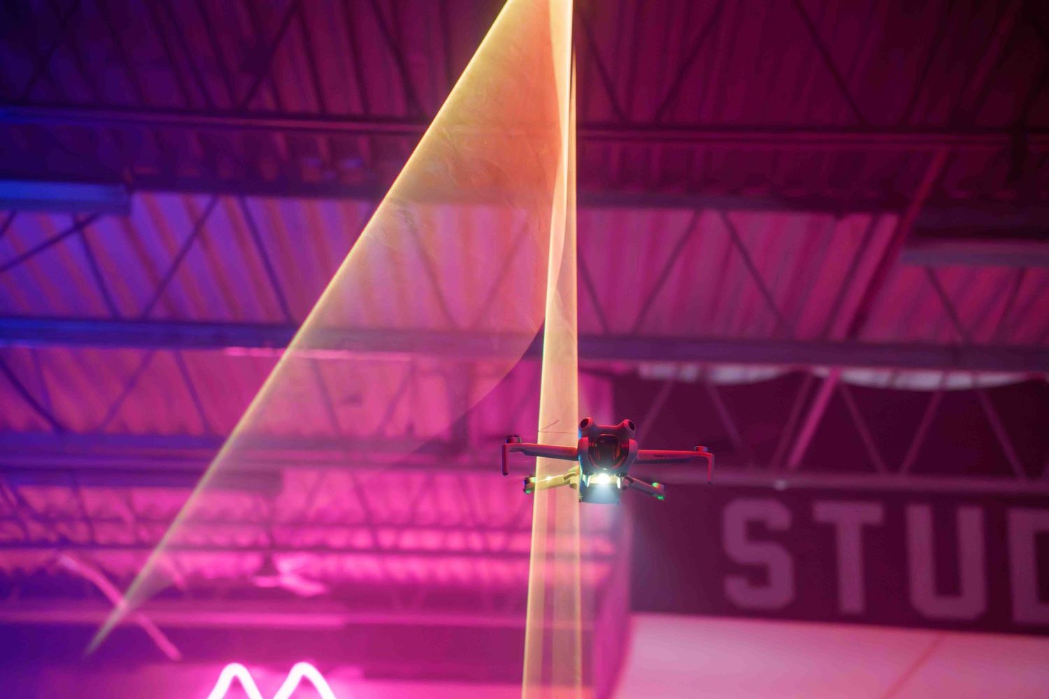 A drone flies through colorful laser beams in a studio with pink and purple lighting.