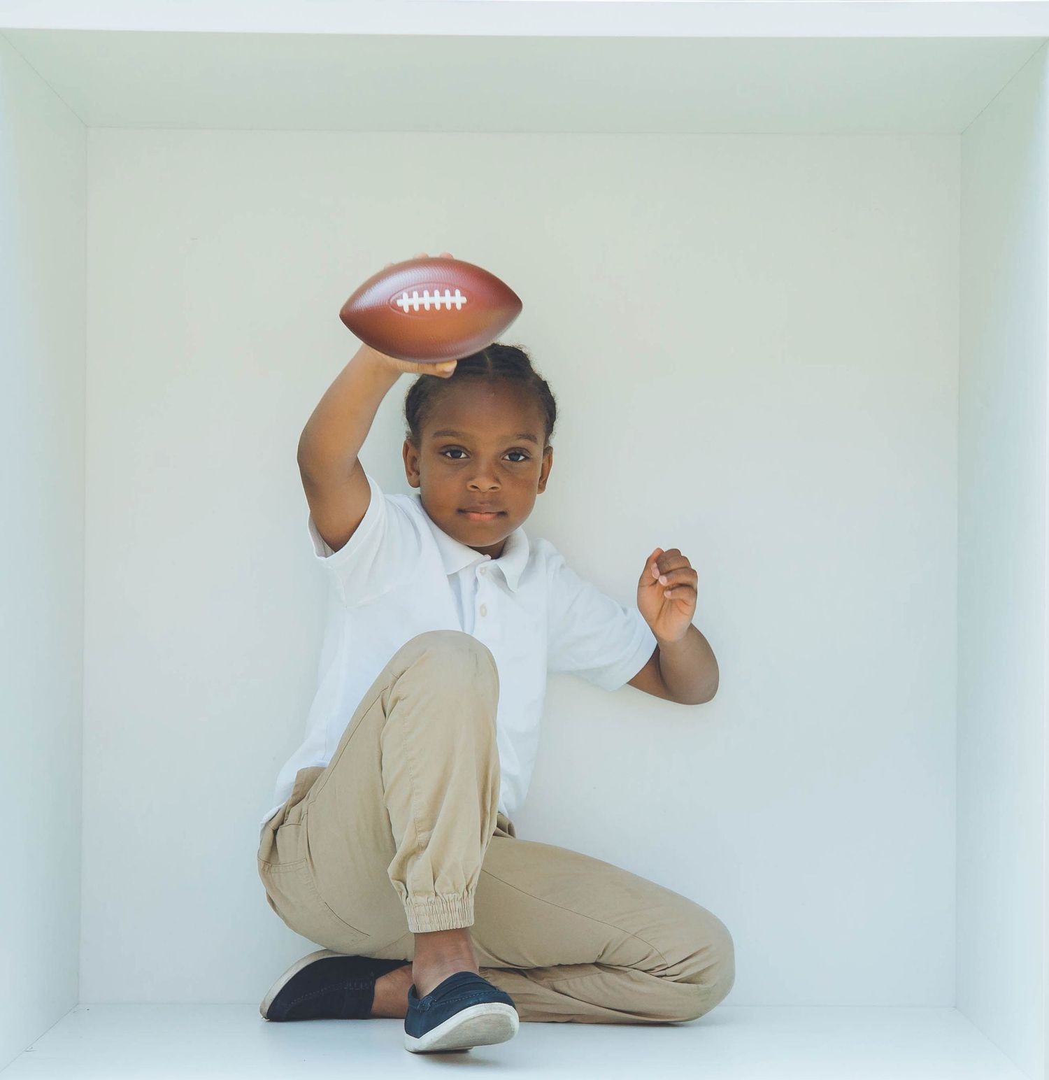 Young child in khaki pants and white shirt playing with a football in a white studio setting.