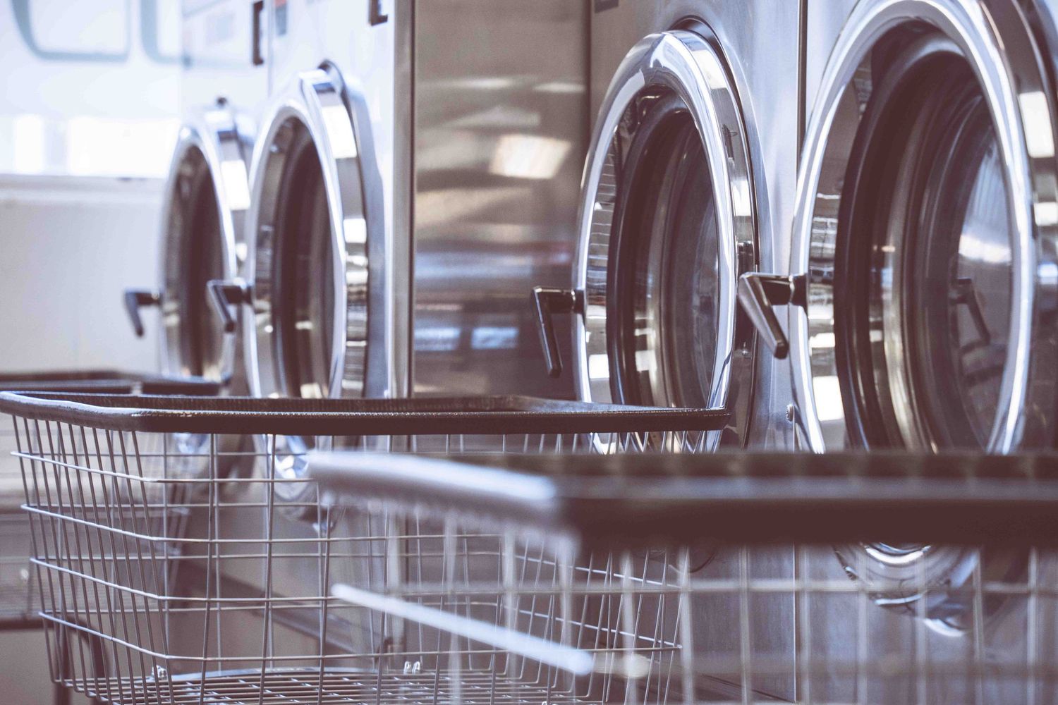 Row of stainless steel front-load washing machines in a laundromat with a wire laundry basket in the foreground.