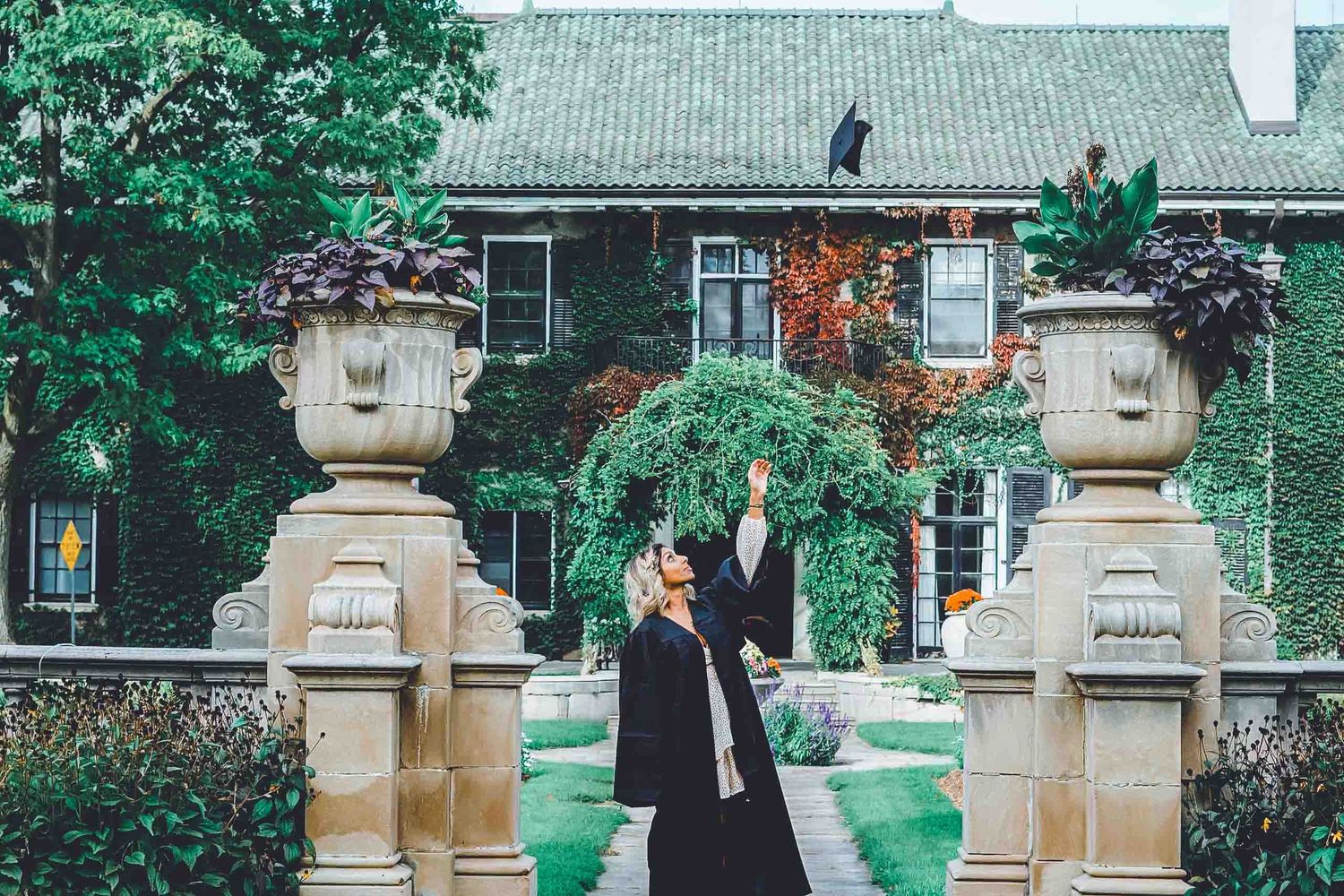 Graduate posing near stone columns and greenery on campus grounds.