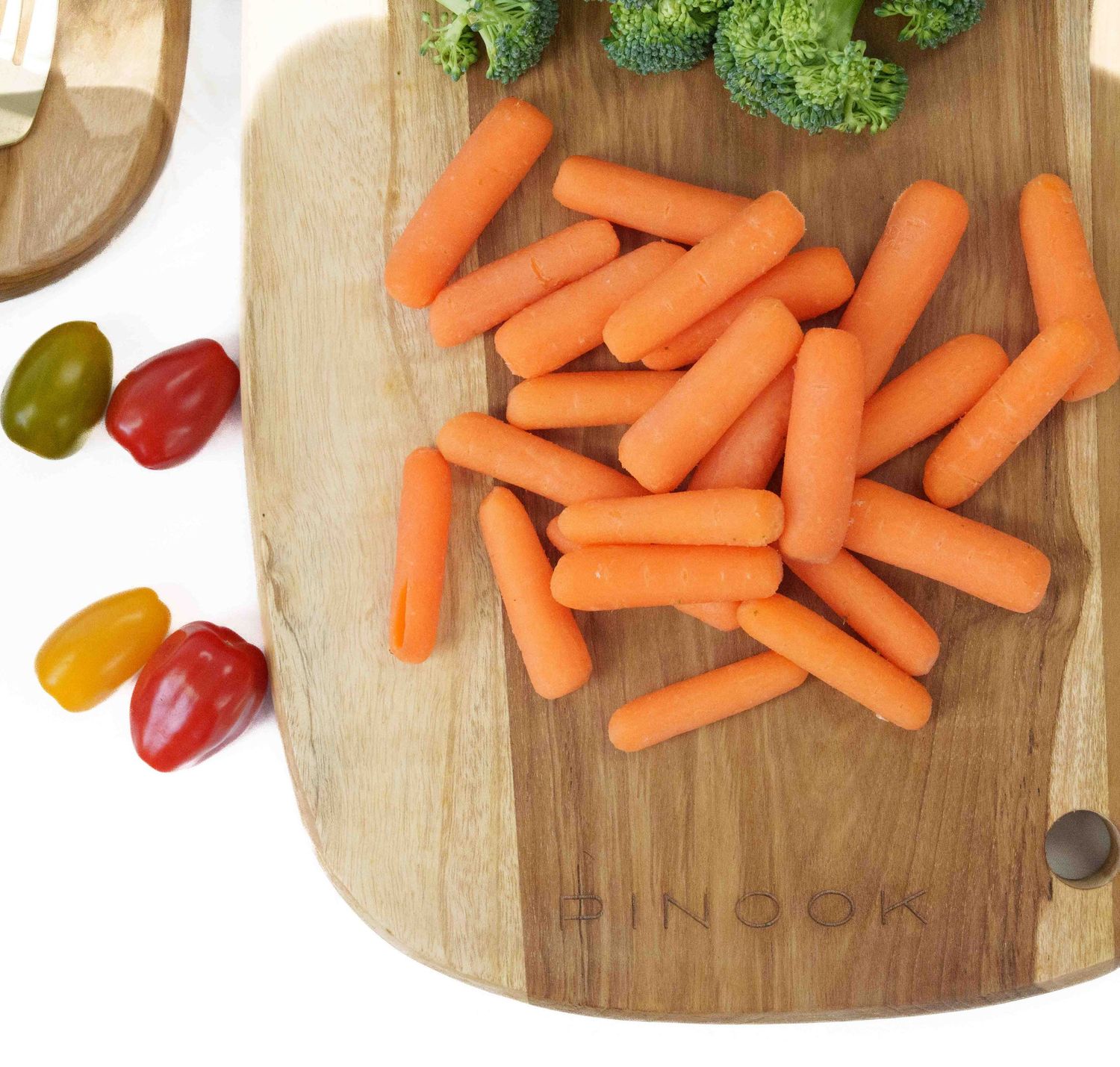 Baby carrots piled on a Pinook wooden cutting board with broccoli and cherry tomatoes nearby.
