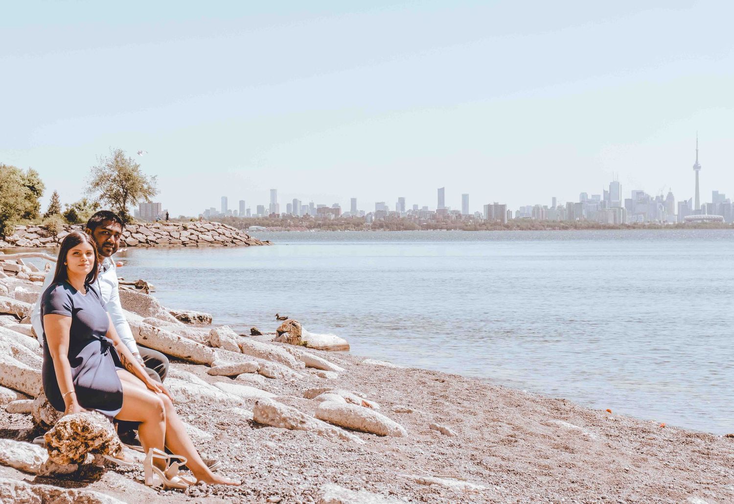 Scenic beach landscape with rocky shoreline and distant city skyline under bright sky.