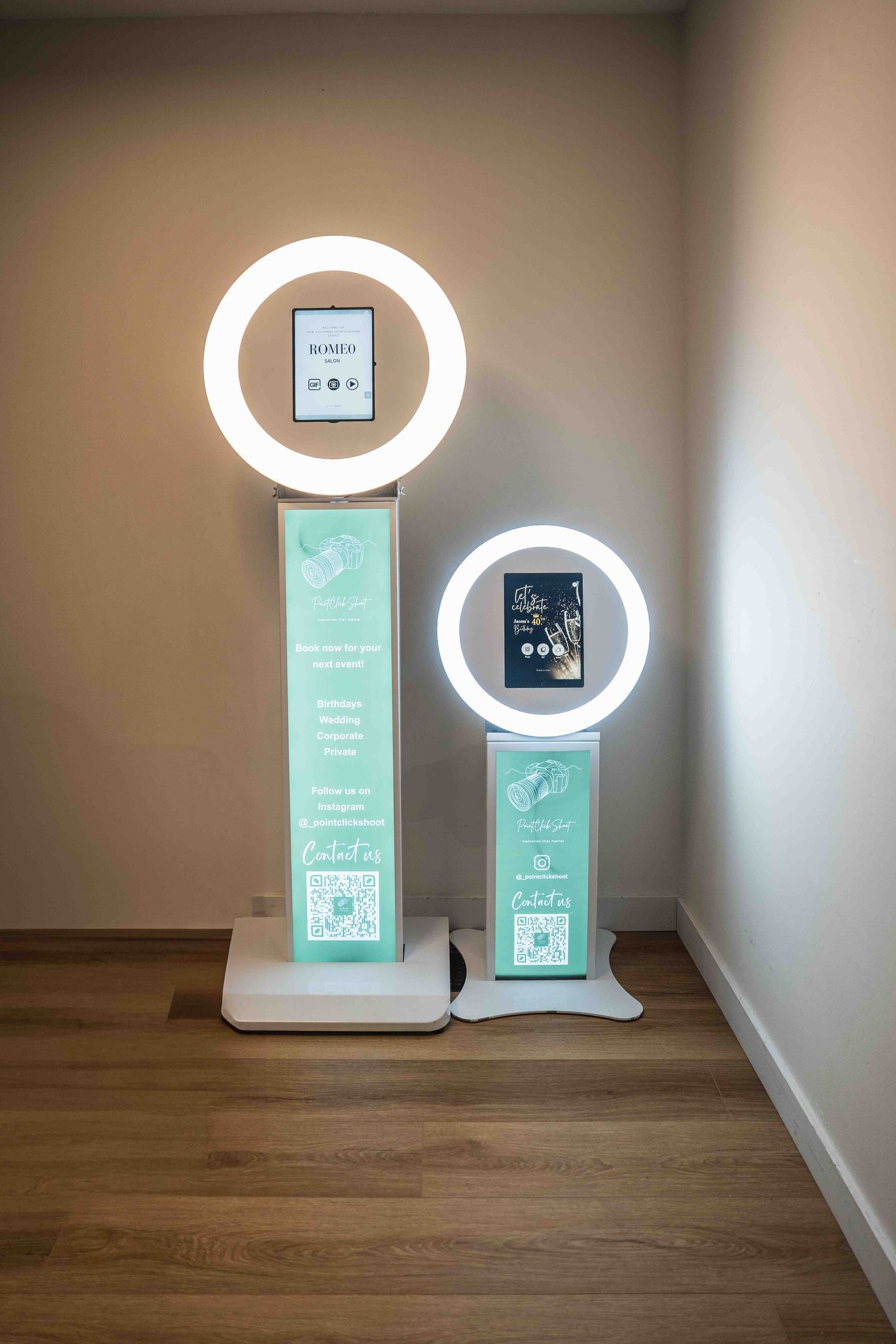 Photo booth setup against white wall with wooden flooring showing digital displays and ring lights.
