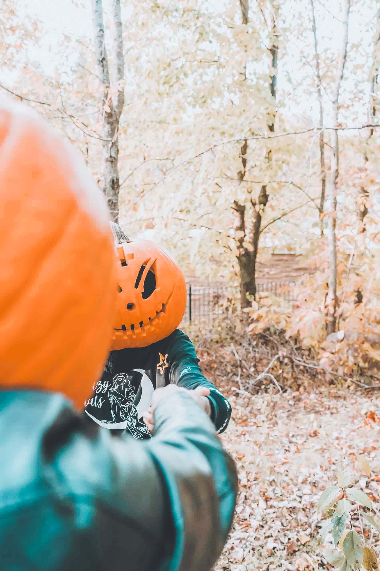 Someone wearing a pumpkin mask and teal jacket photographs in an autumn forest.