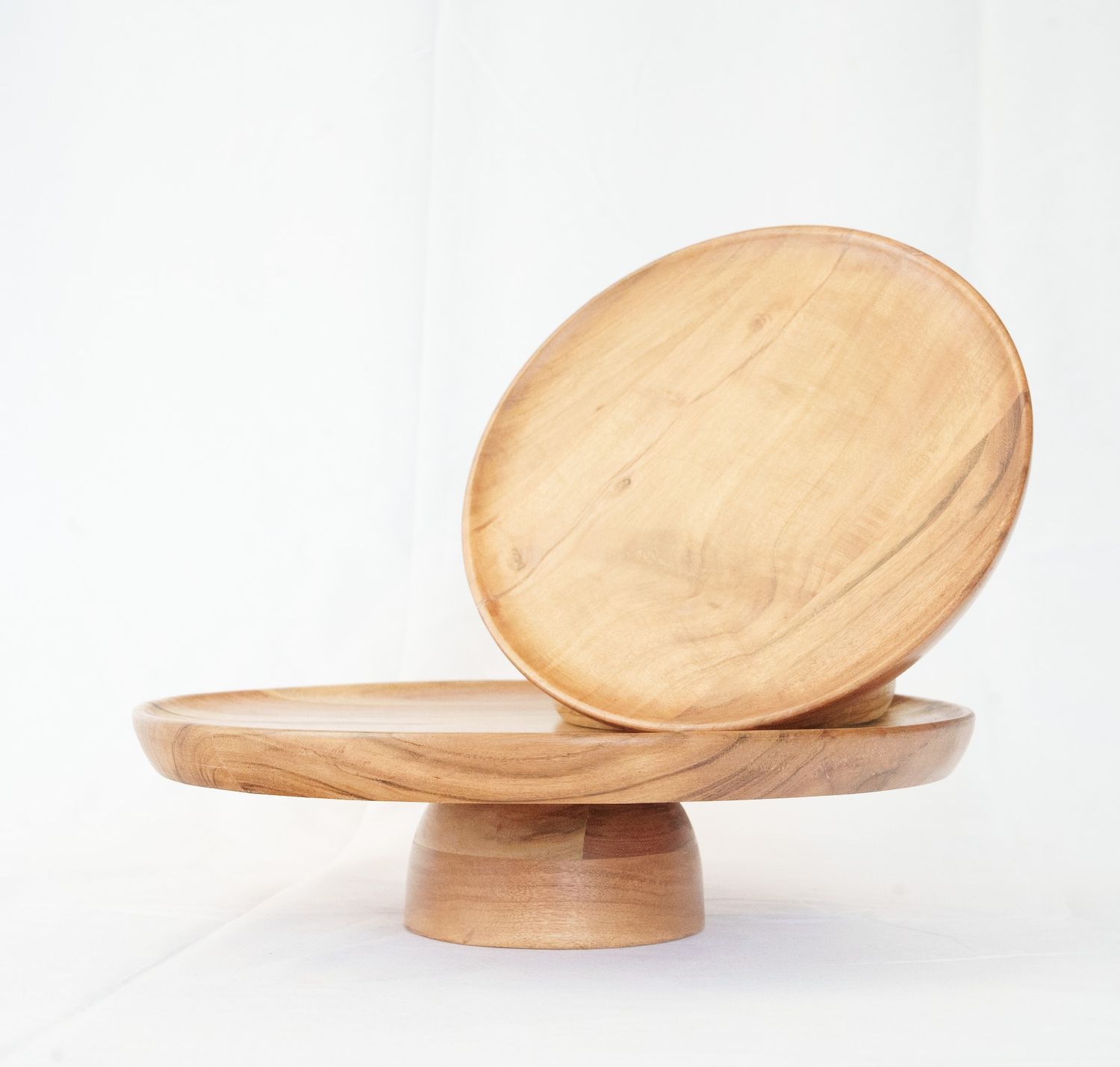 Two round wooden cake stands with pedestal bases, one upright and one leaning, showcasing natural wood grain finish.