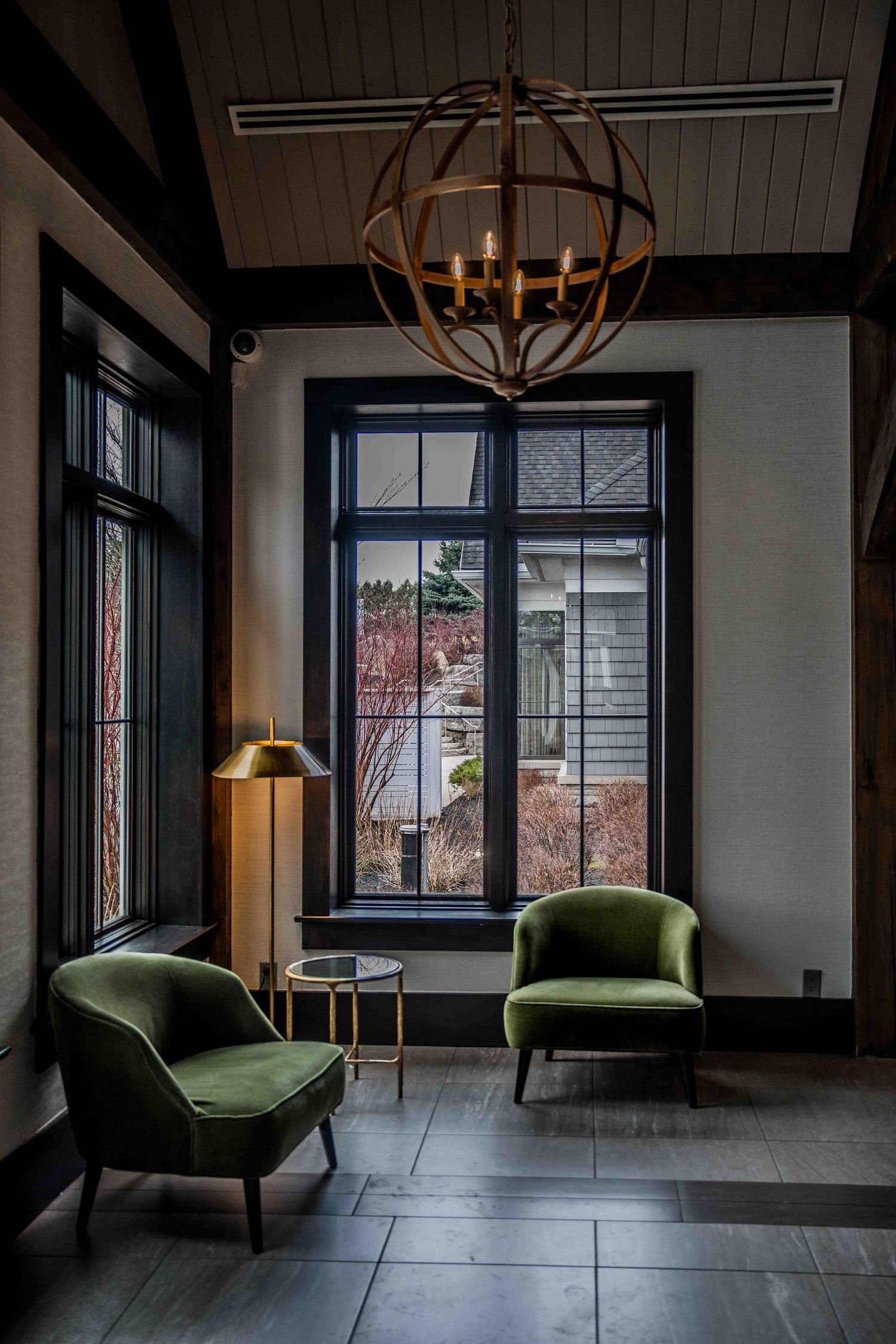 Elegant sitting area with green velvet chairs, gold chandelier, tall black-framed windows, and a brass floor lamp on slate tile flooring.