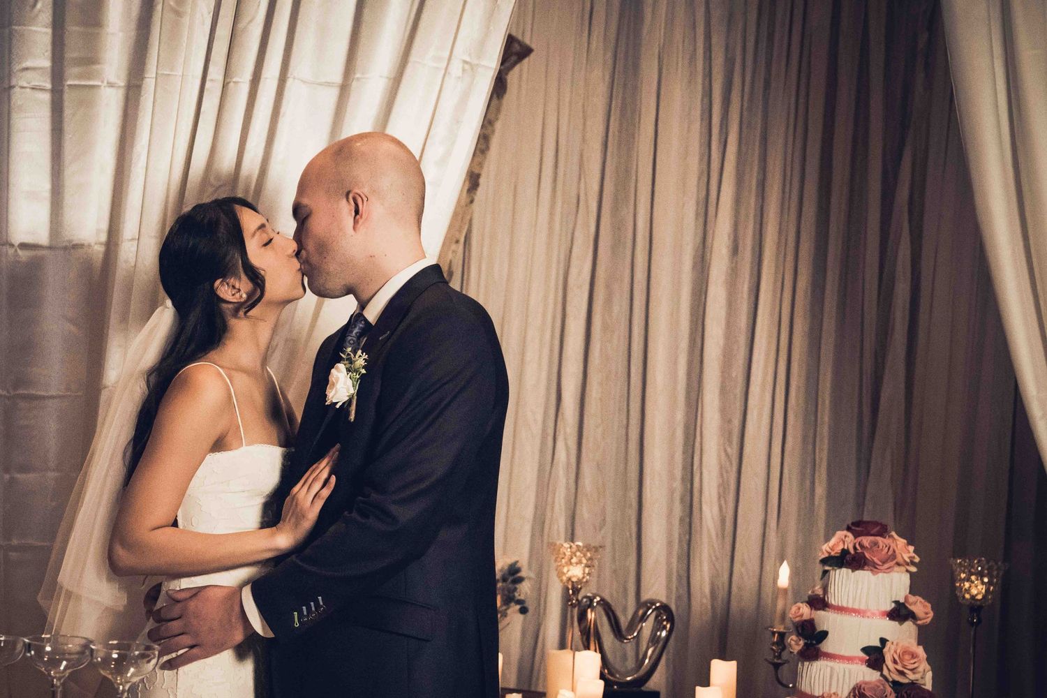 Romantic wedding couple shares intimate moments in elegant indoor venue with soft lighting and flowing curtains.