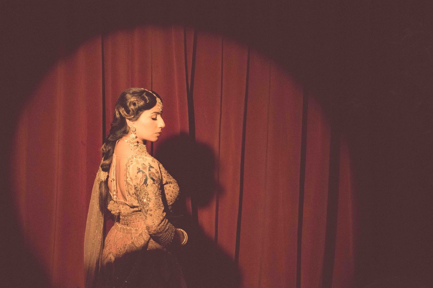 Dramatic portrait of a bride in vintage attire illuminated by a spotlight against red curtains.
