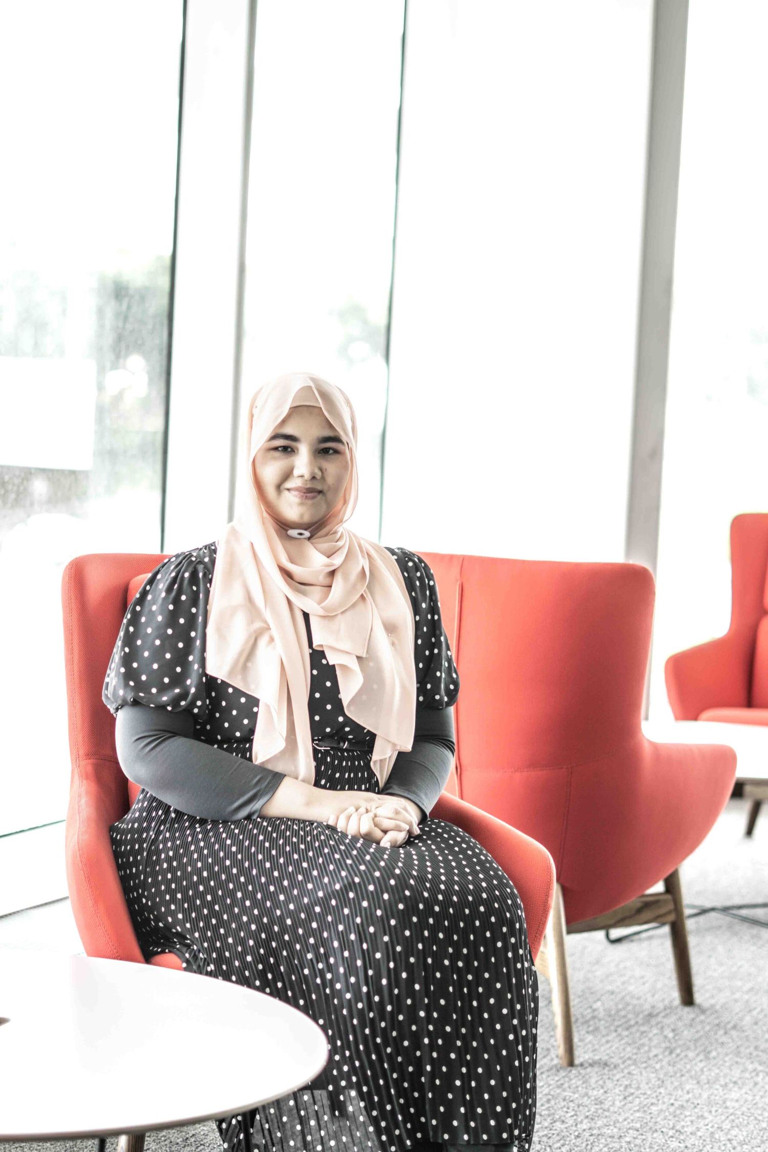 Person in polka dot dress and light headscarf seated on red chair in bright modern space.
