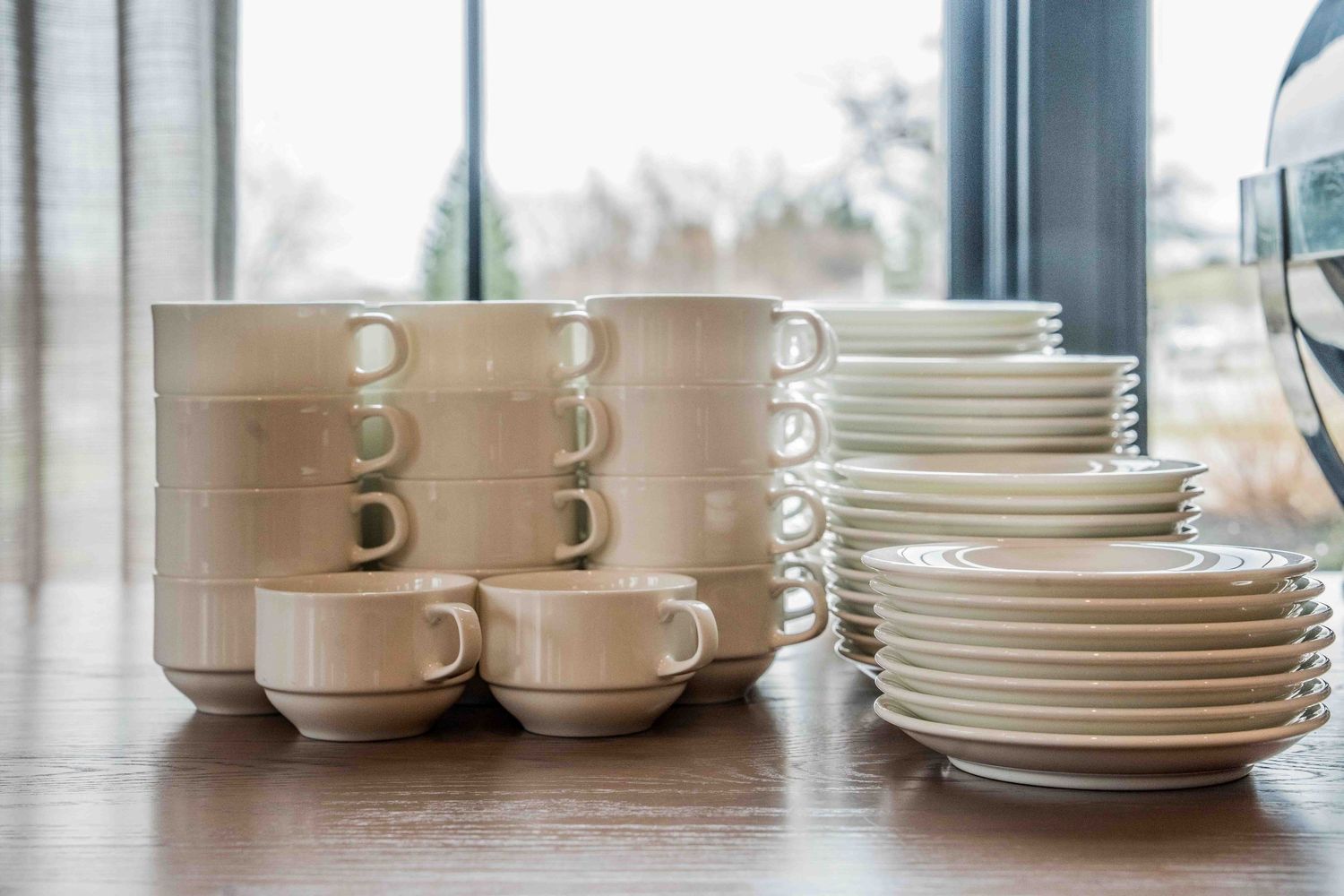 Stacked white ceramic cups, saucers, and plates arranged neatly on a wooden table near a window.