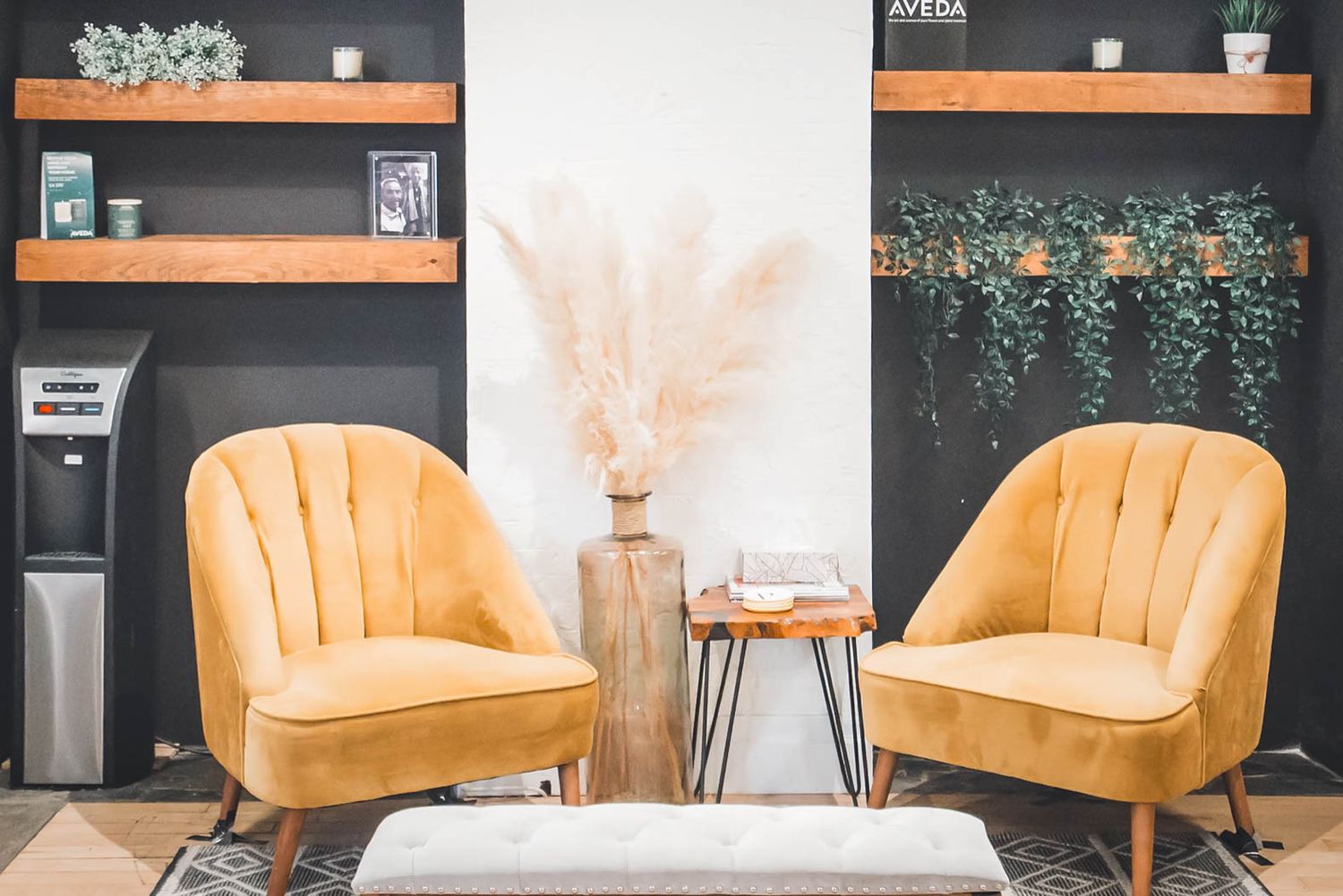Two golden velvet accent chairs flank a wooden vase with pampas grass in a modern salon waiting area.