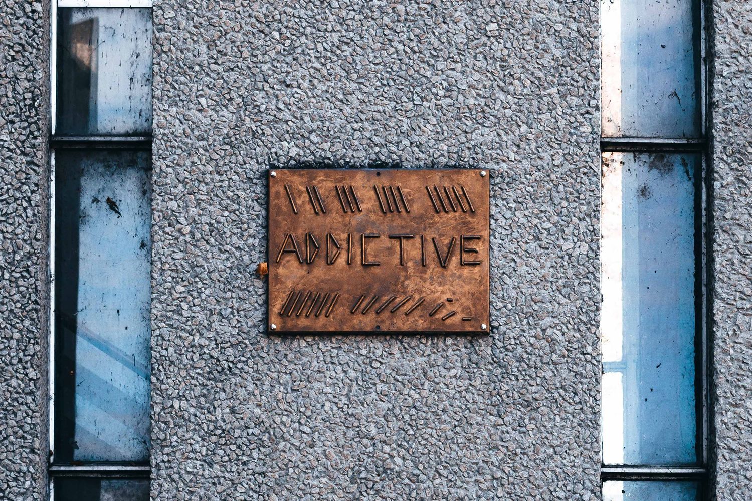 Weathered metal sign reading 'Addictive' mounted on textured concrete wall between two windows.