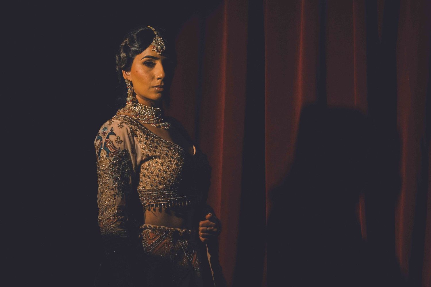 A bride in an ornate traditional Indian wedding dress poses in dramatic side lighting against red curtains.