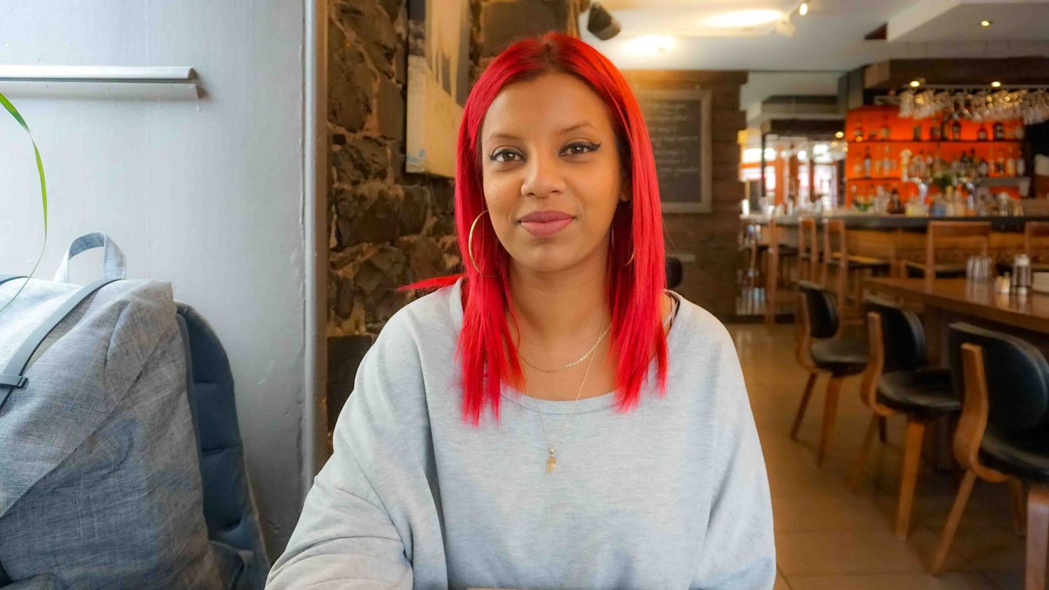 Person with vibrant red hair wearing gray sweatshirt seated in casual restaurant setting.