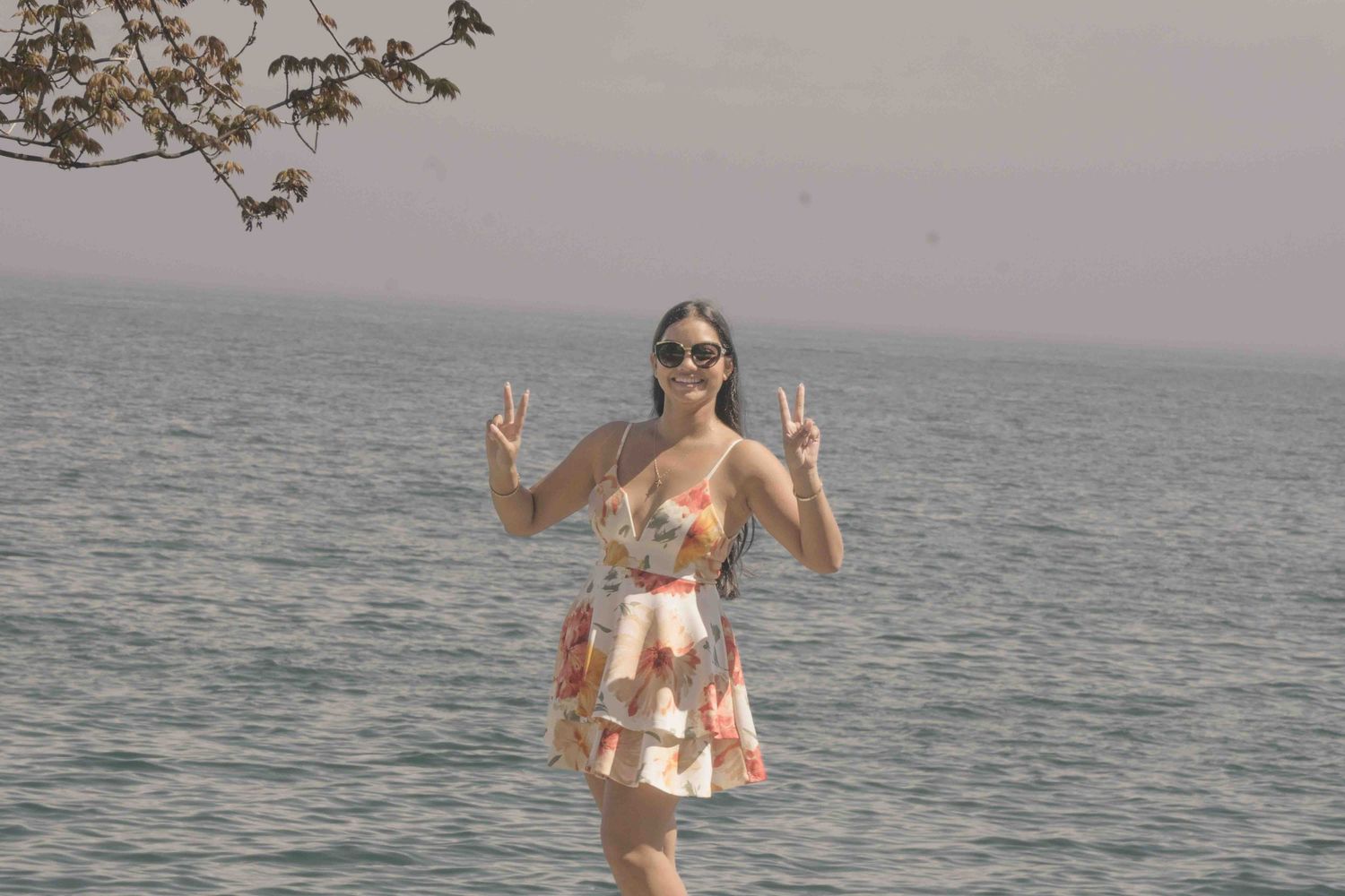 A person in a floral dress makes peace signs while posing by the ocean.