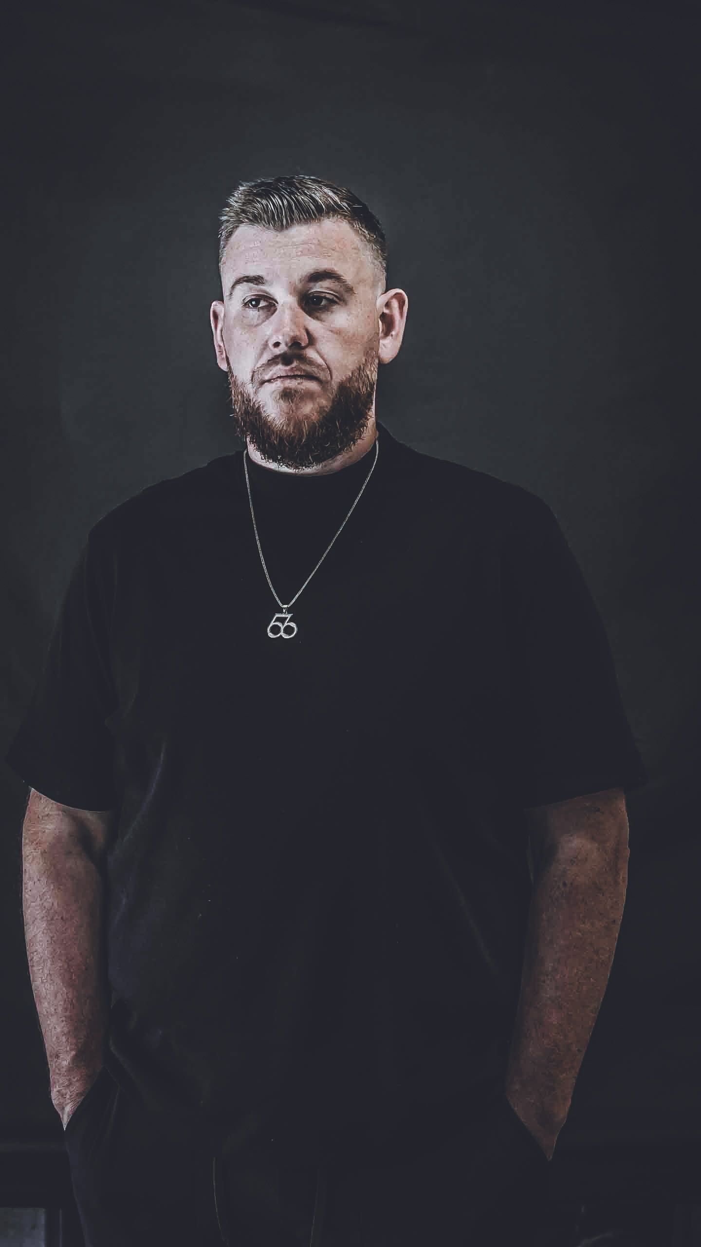 Professional portrait of person in black t-shirt with necklace against dark background.