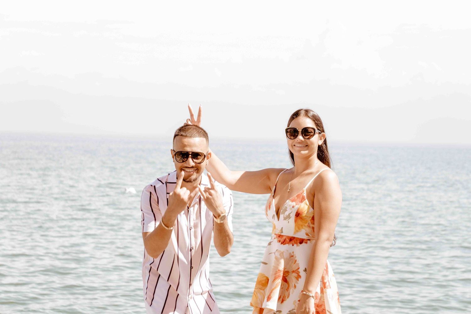 A playful couple poses by the ocean, making peace signs and wearing summer attire.