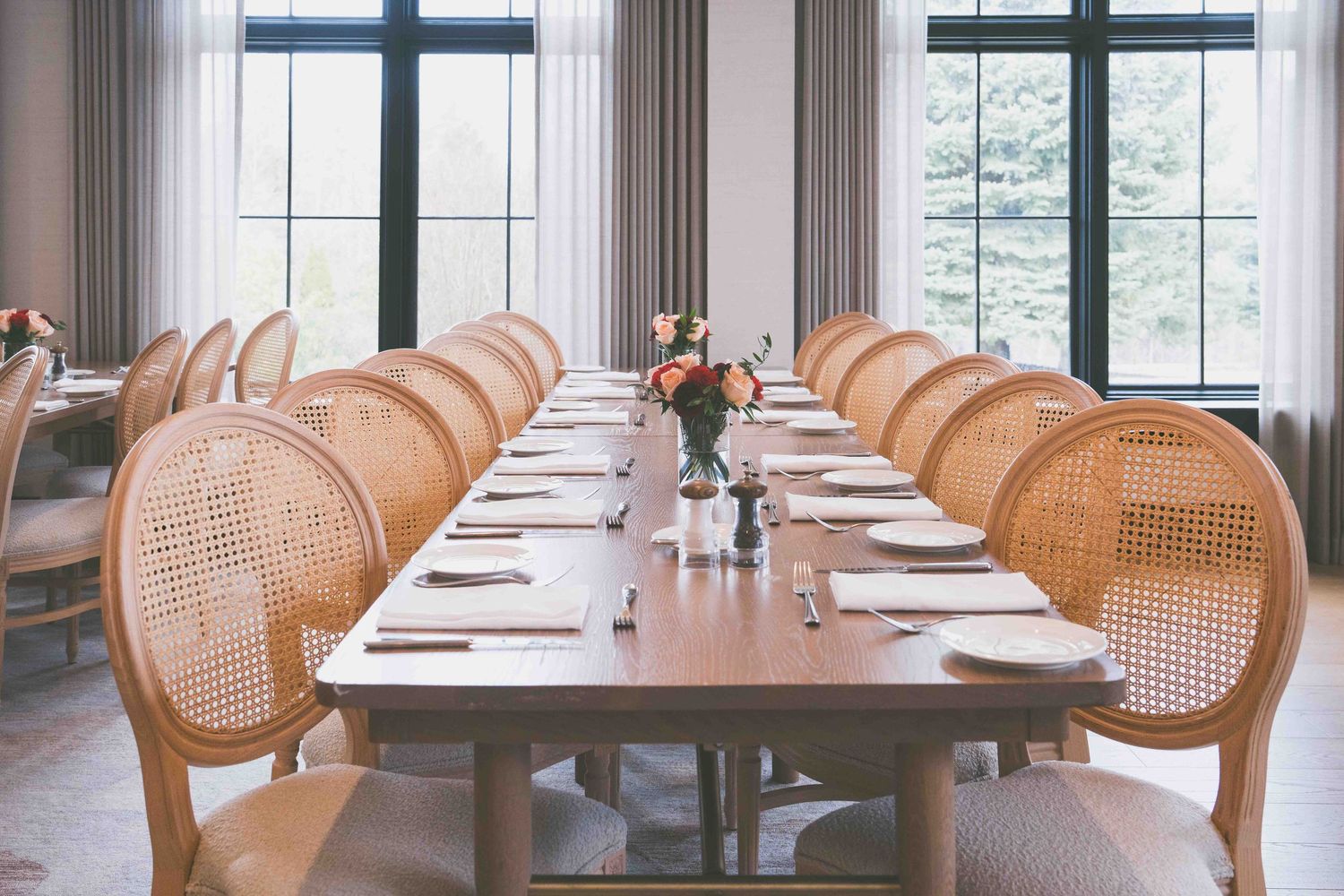 Elegant dining room with long wooden table, woven rattan chairs, floral centerpiece, and large windows with sheer curtains.