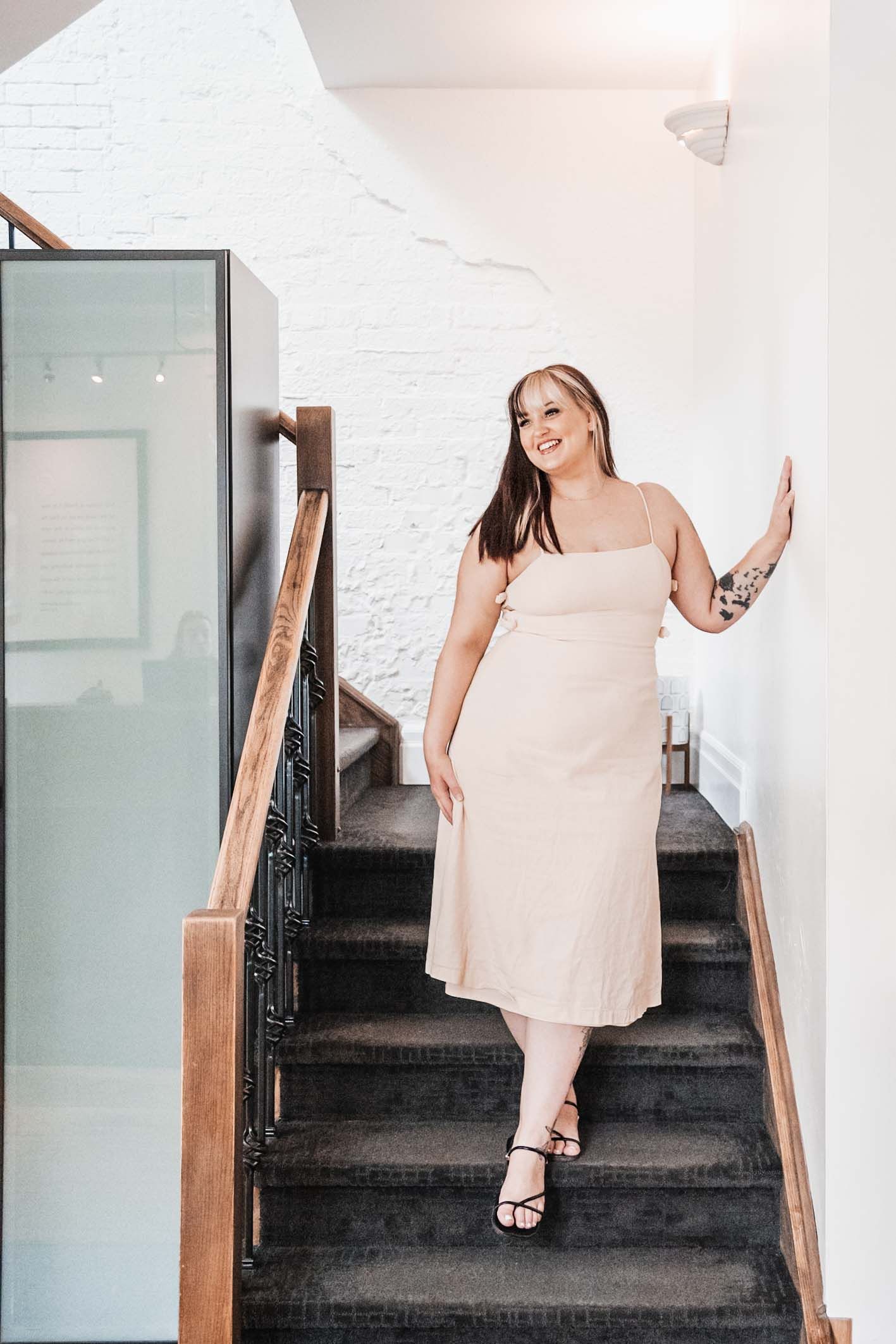 A person in a light dress poses on dark wooden stairs in a bright interior space.