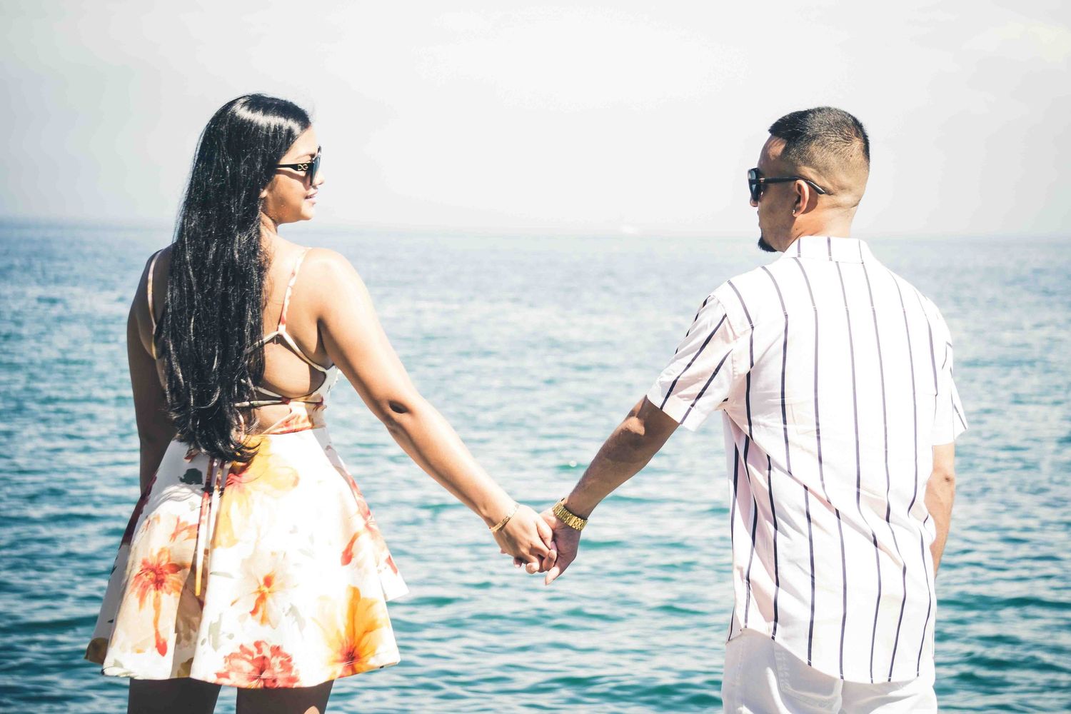 A couple holds hands while looking out at the ocean, wearing coordinated summer outfits.
