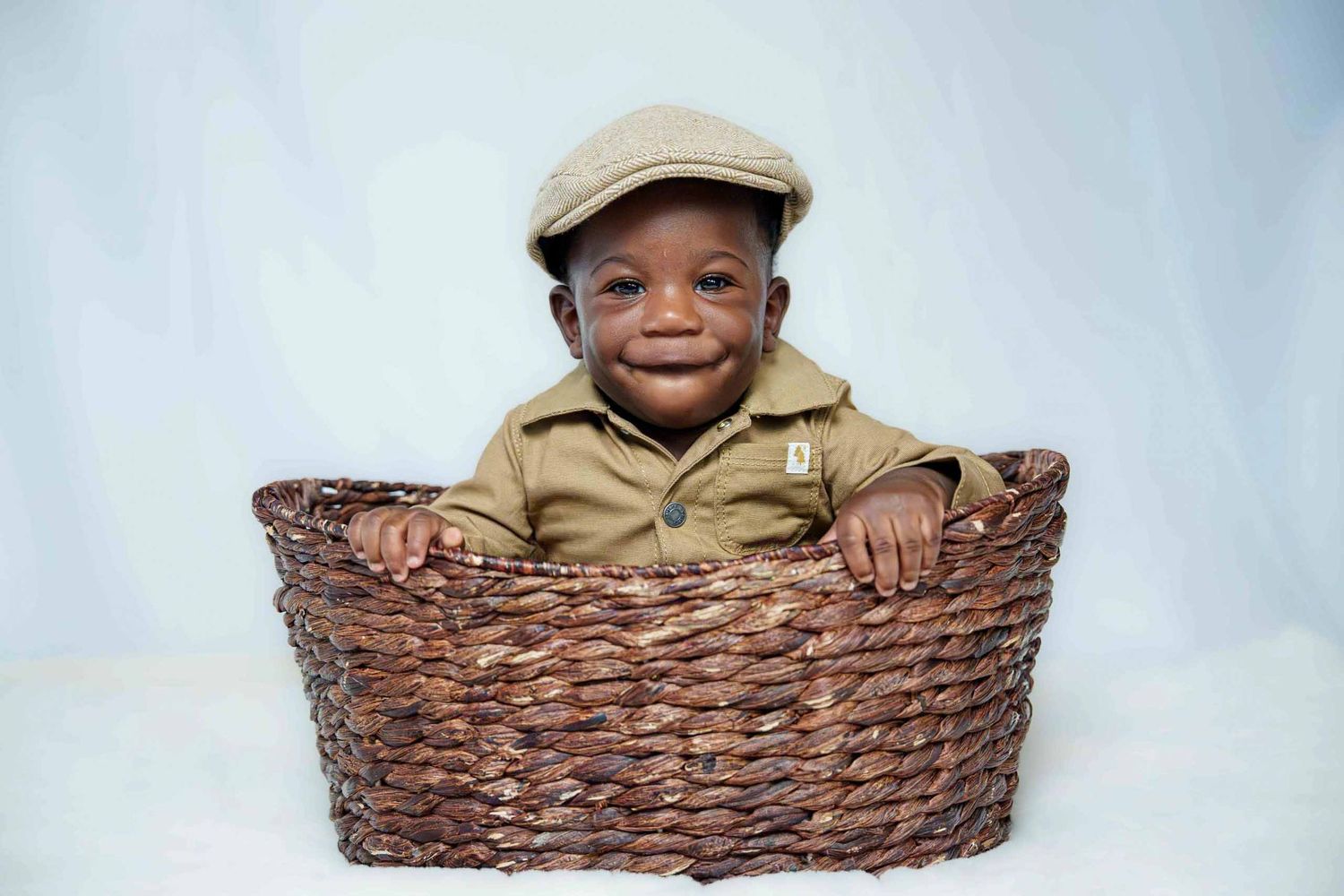 Child in tan outfit and cap sitting cheerfully in woven basket.
