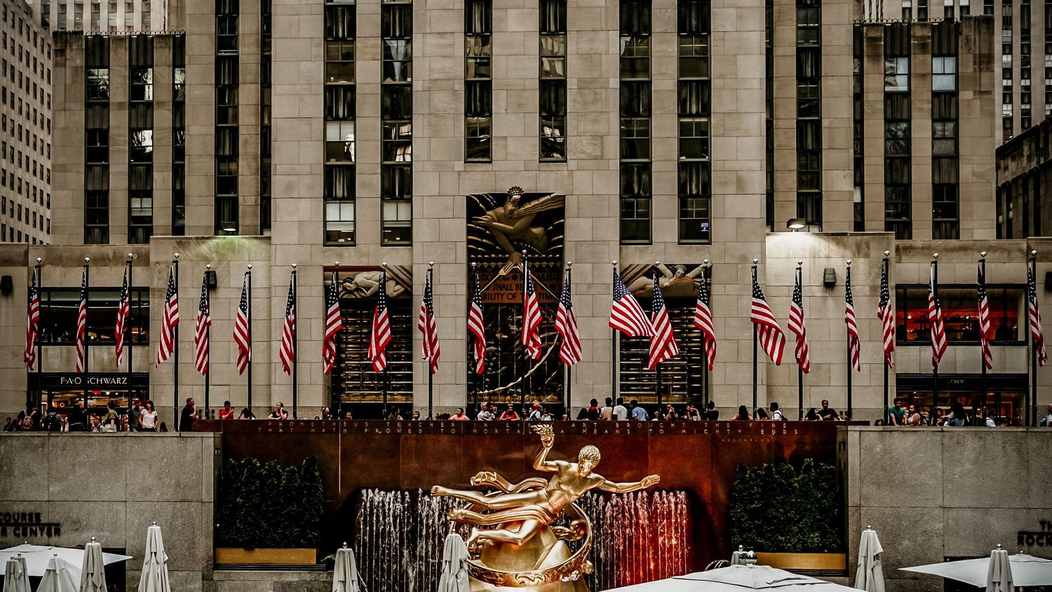 Wide angle view of Rockefeller Center plaza with American flags and Art Deco architecture.