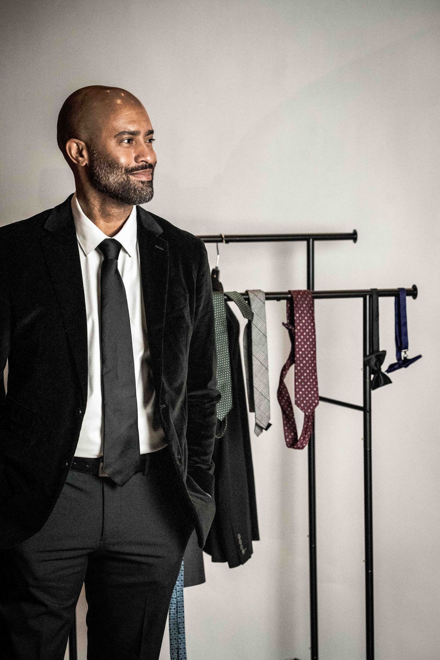 Person in black suit standing by clothing rack with colorful ties against light background.
