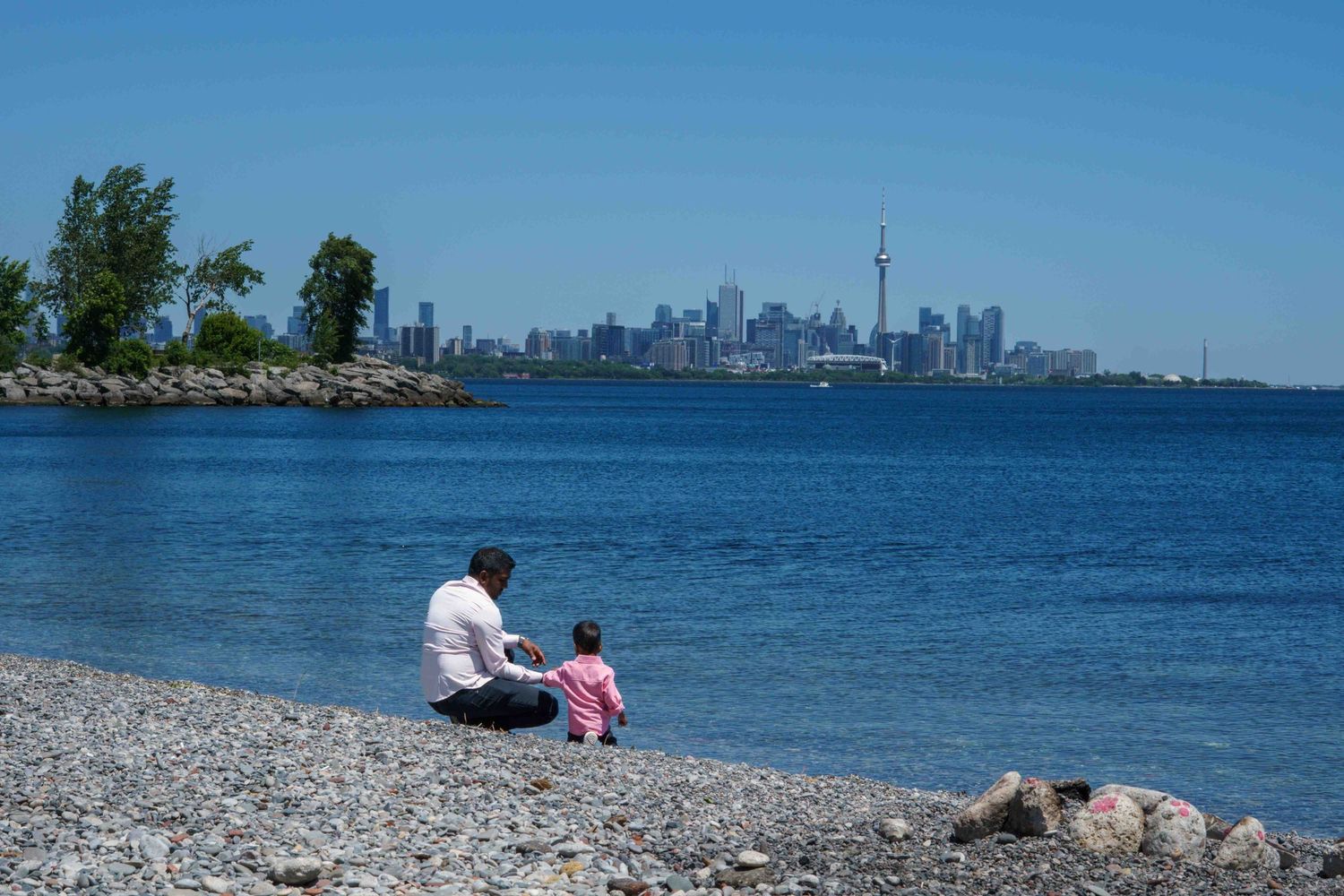 Rocky beach shoreline with Toronto city skyline visible in the distance on a clear sunny day.