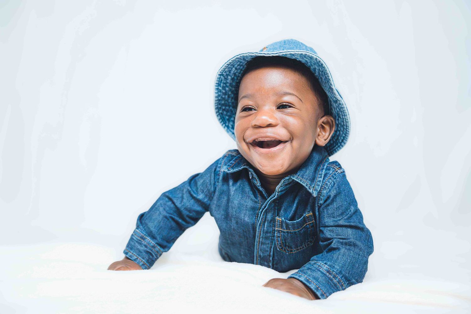 Baby wearing blue denim hat lying on white background showing joyful expressions.