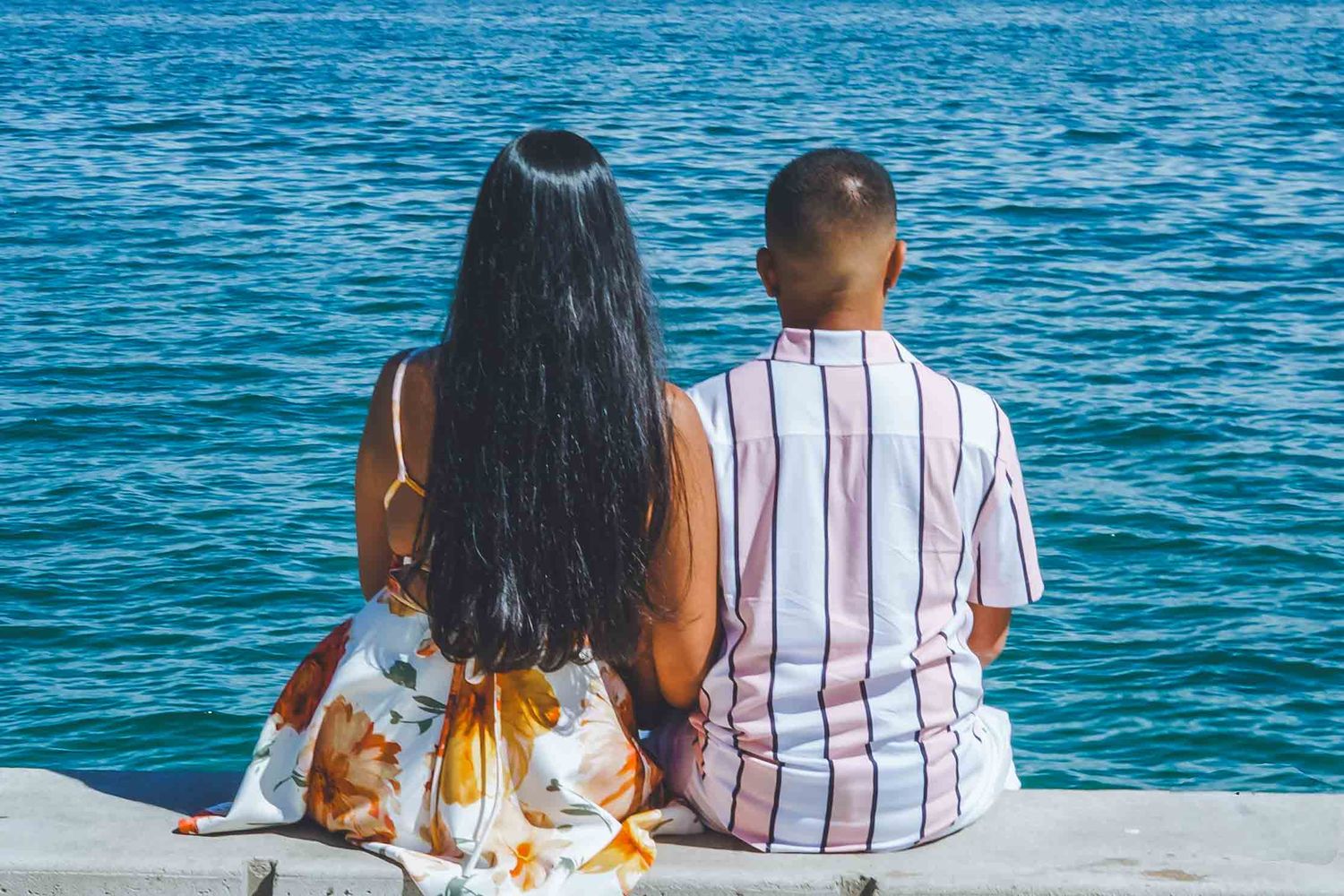 Couple sitting together on dock overlooking bright blue ocean waters.