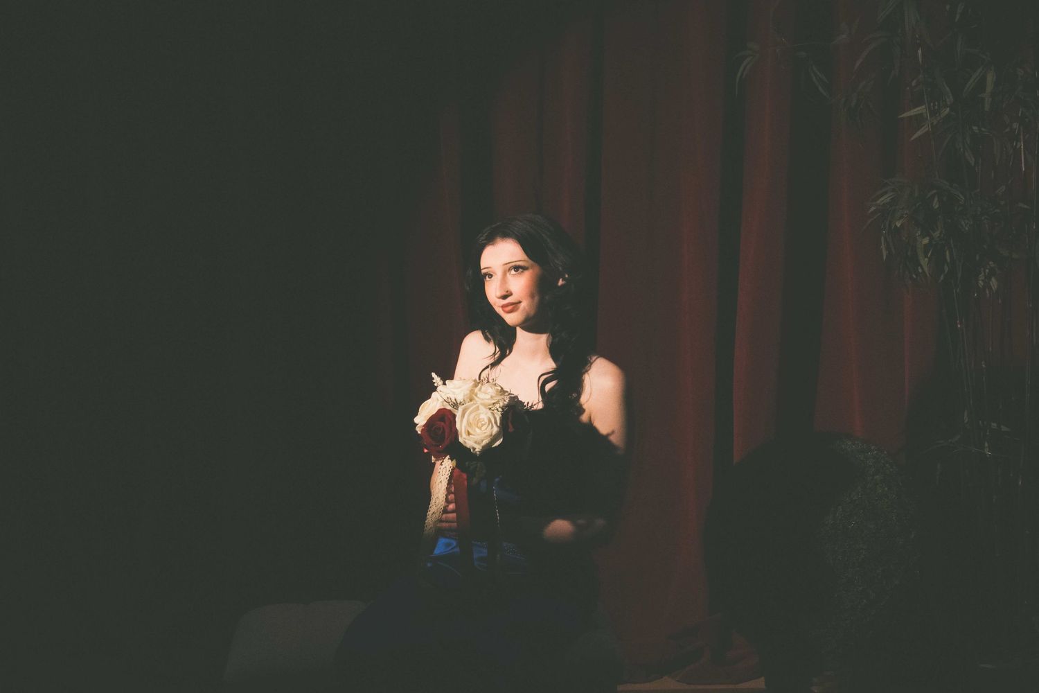 A performer in a black dress holds a microphone while singing on a dimly lit stage with red curtains in the background.