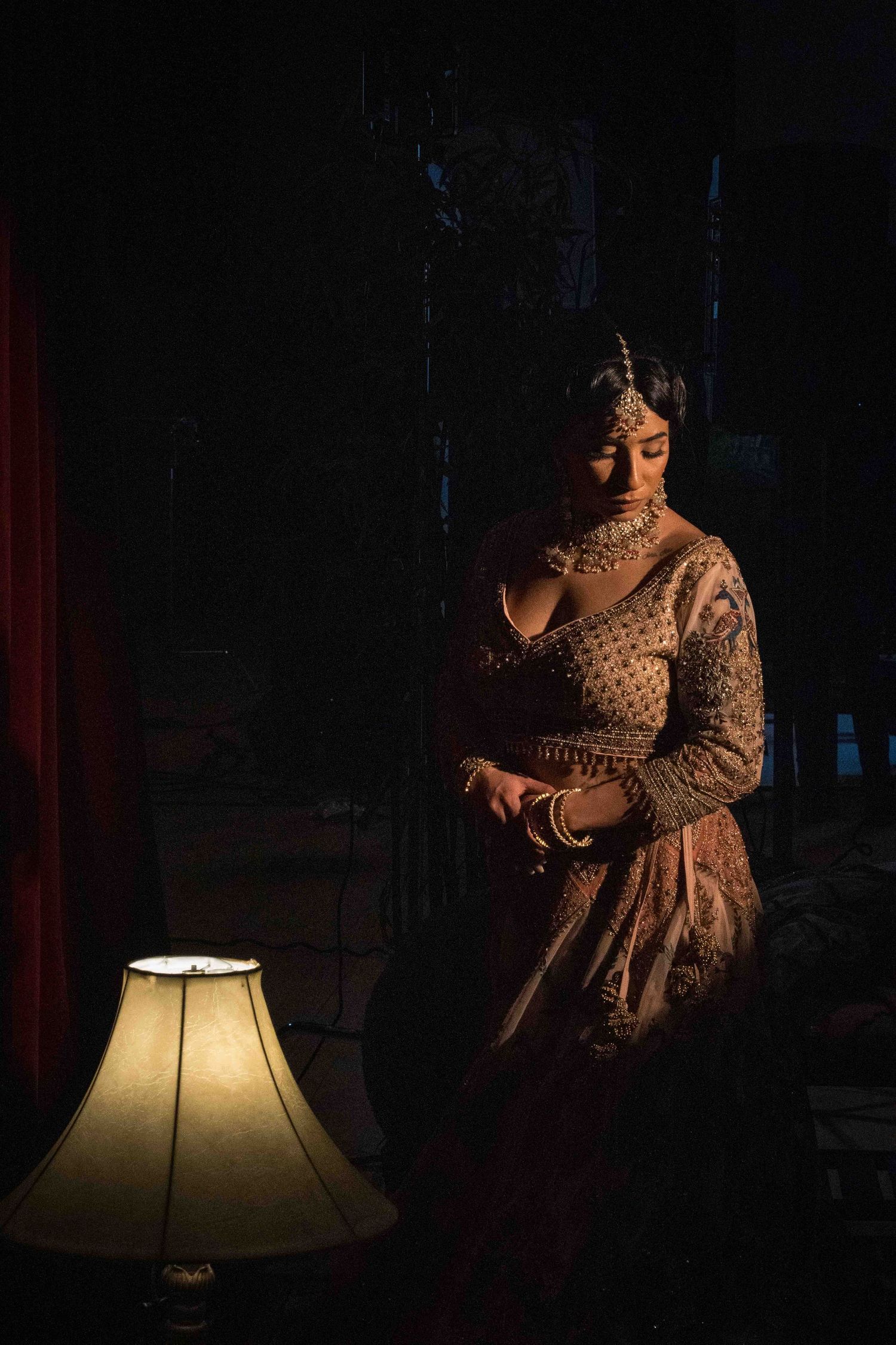 A person in traditional Indian attire stands in dramatic lighting next to an illuminated table lamp in a dark room.