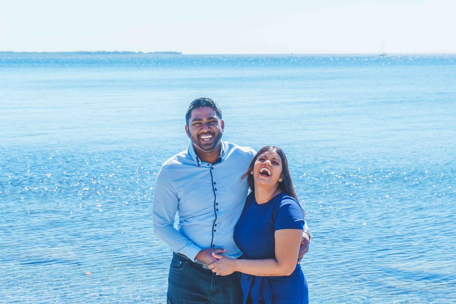 Romantic couple embracing on beach with blue water and clear sky in background.