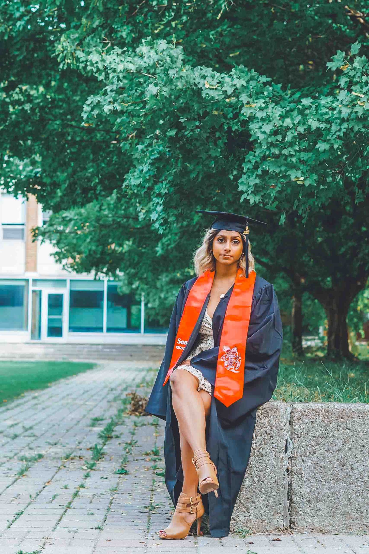 Graduate in cap and gown with orange stole posing on campus pathway.