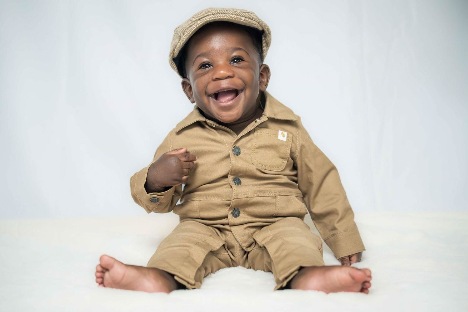 Happy child in khaki outfit and cap sitting in woven basket against white backdrop.